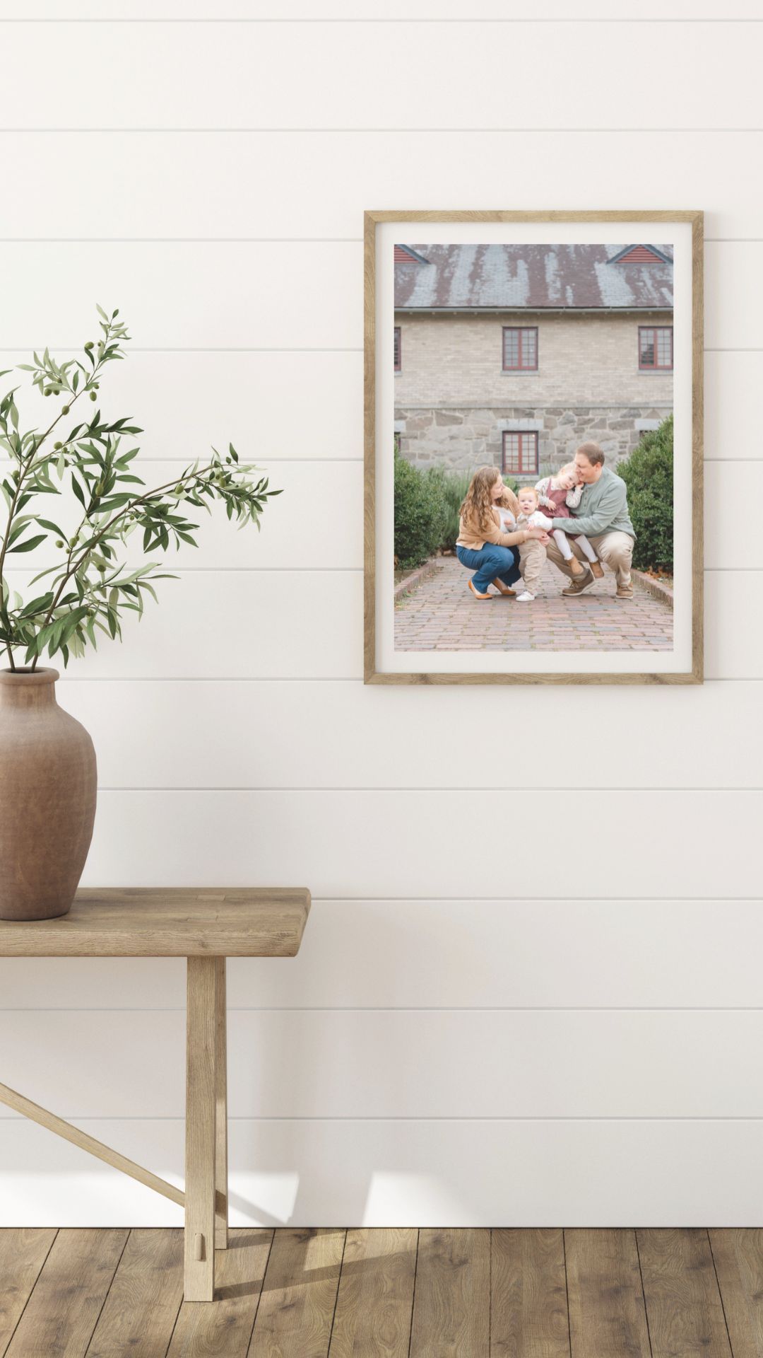 Framed family photo in wood frame on white shiplap wall above rustic bench with terracotta vase and greenery.