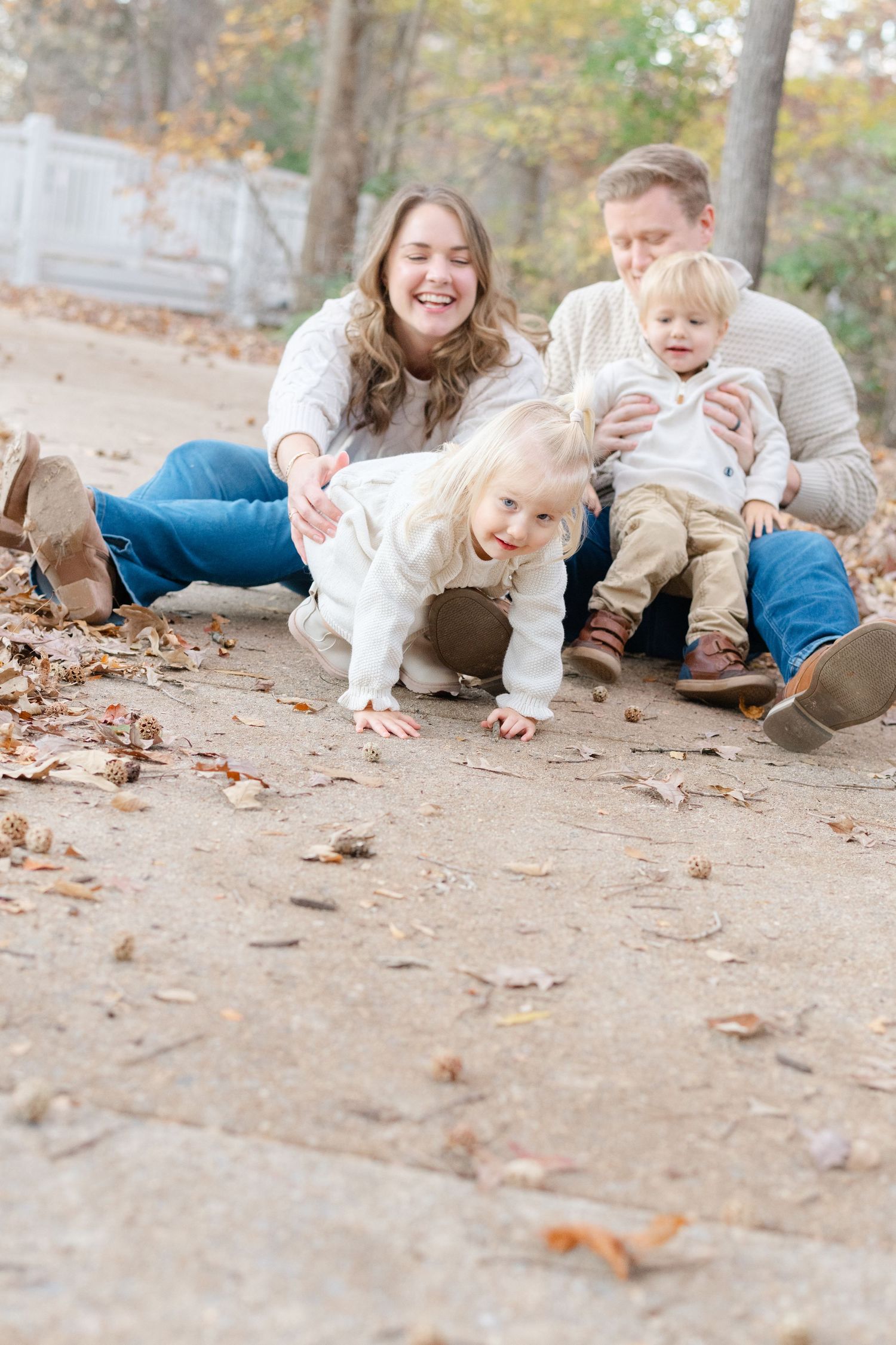 Family of four enjoying a playful autumn day outdoors, with toddler crawling and parents laughing on a leaf-covered path.