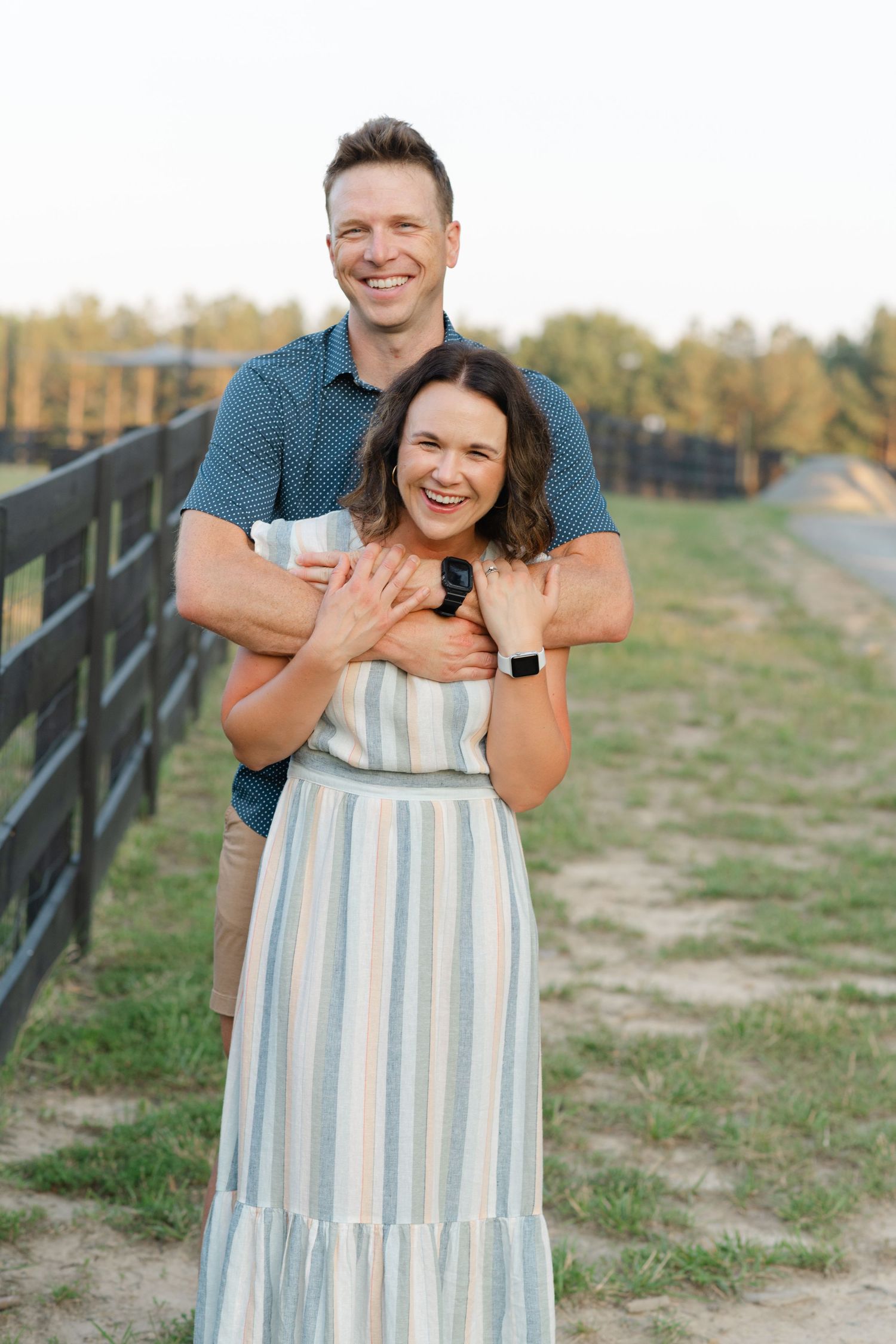 A smiling couple embraces outdoors near a black fence, woman in striped maxi dress, man in blue shirt, on a sunny day.