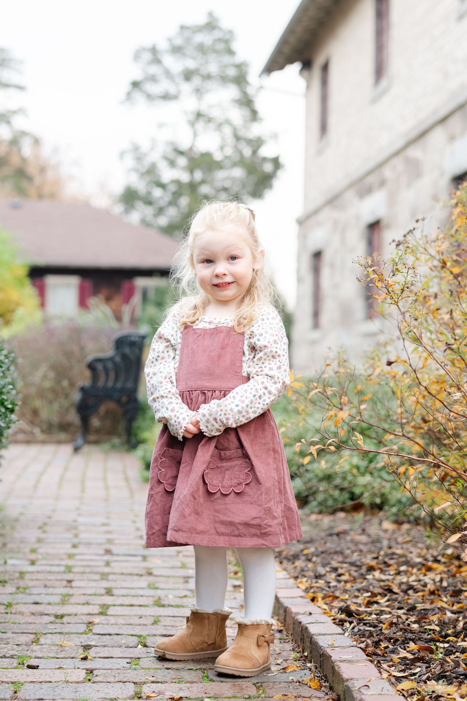 Adorable blonde toddler girl in pink pinafore dress and boots standing on a garden path outdoors.