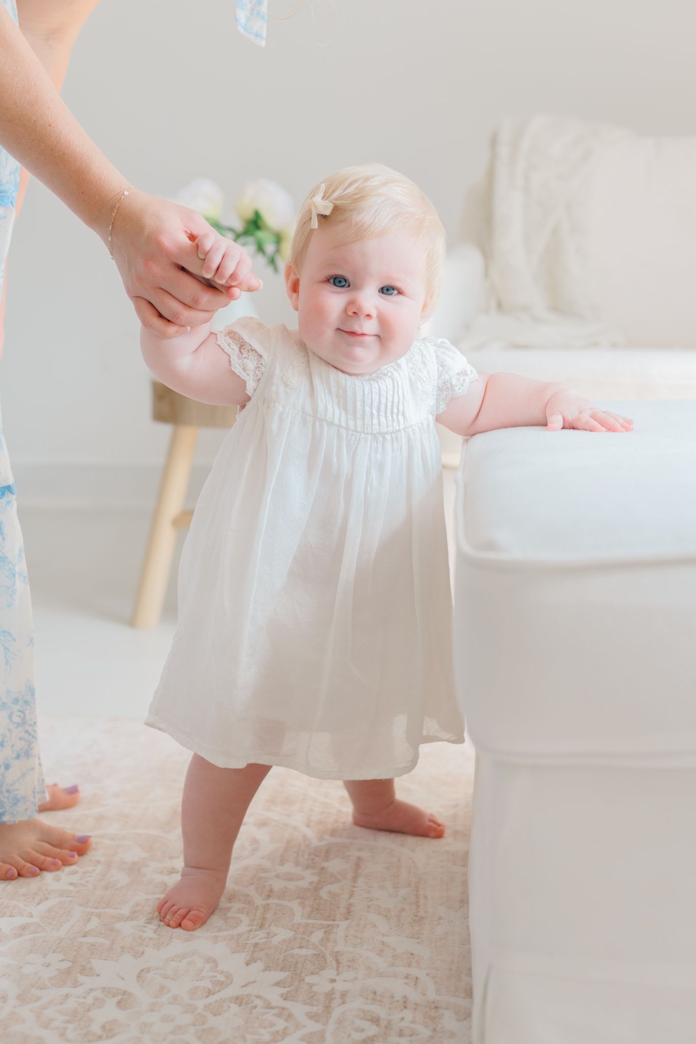 Small child in a white dress takes first steps on a light carpet.