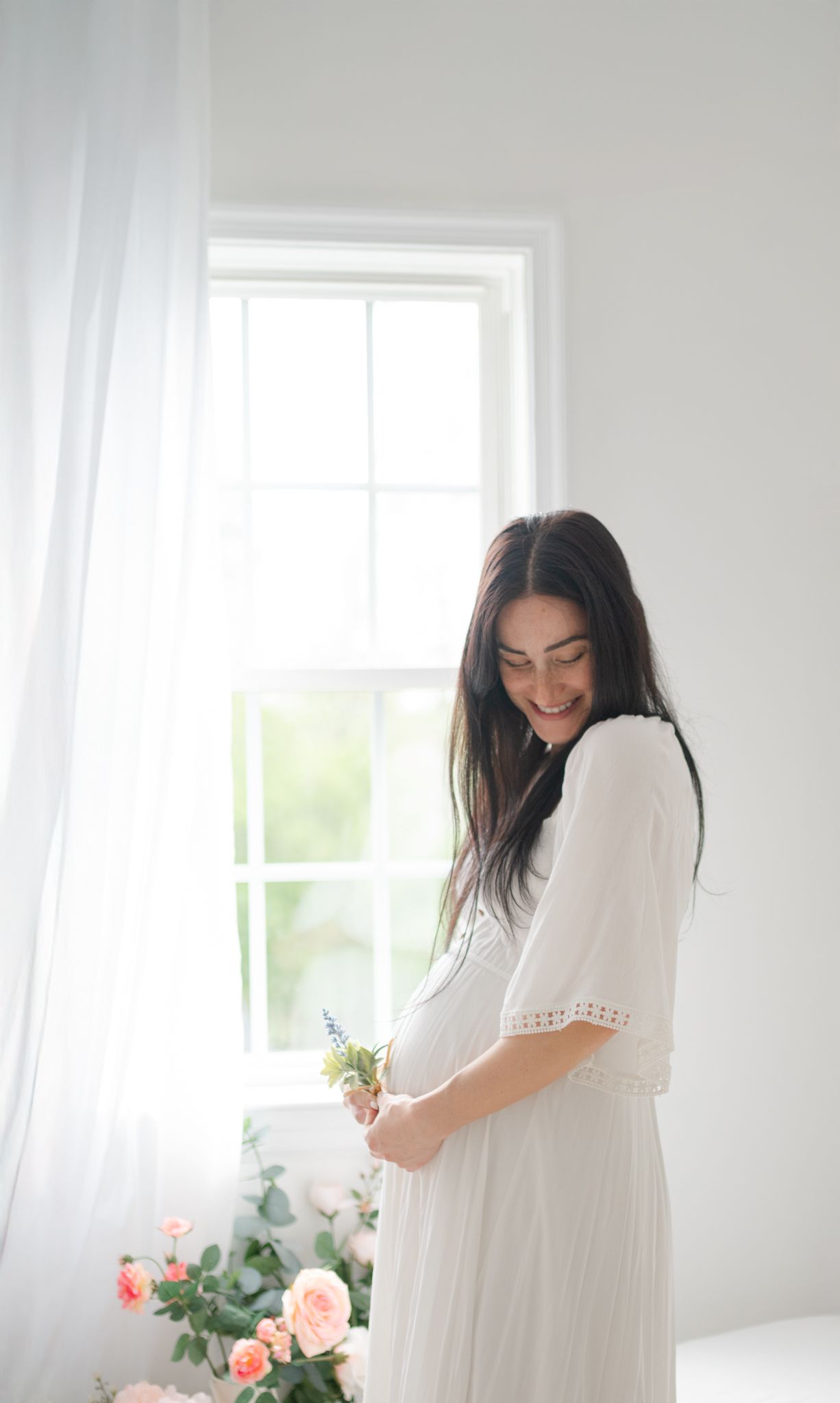 Woman in white dress arranges fresh pink roses in a bright sunlit room with multiple window panels.