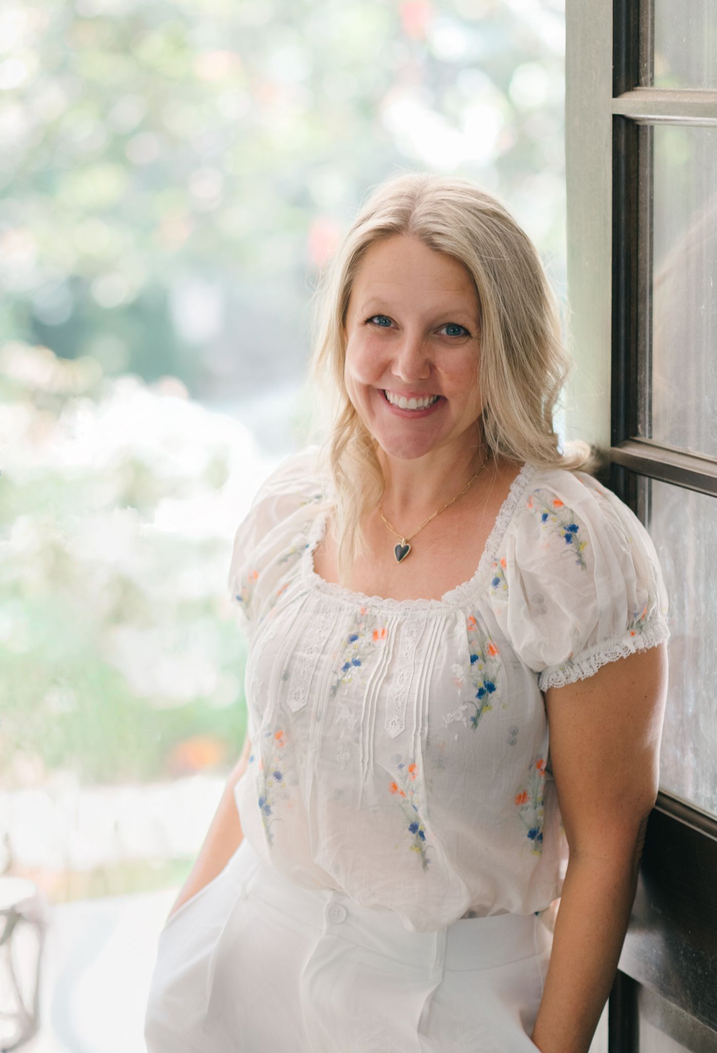 Richmond newborn photographer in a white peasant blouse with floral embroidery smiles while standing near a window in soft natural light.