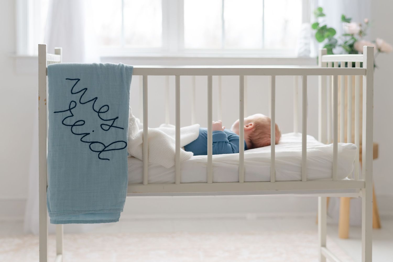 Newborn baby sleeping in white crib with blue personalized knit blanket draped over the side.