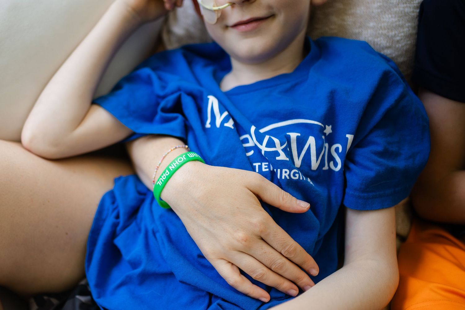 Child wearing a blue Make-A-Wish t-shirt and green wristband, resting comfortably on a couch.
