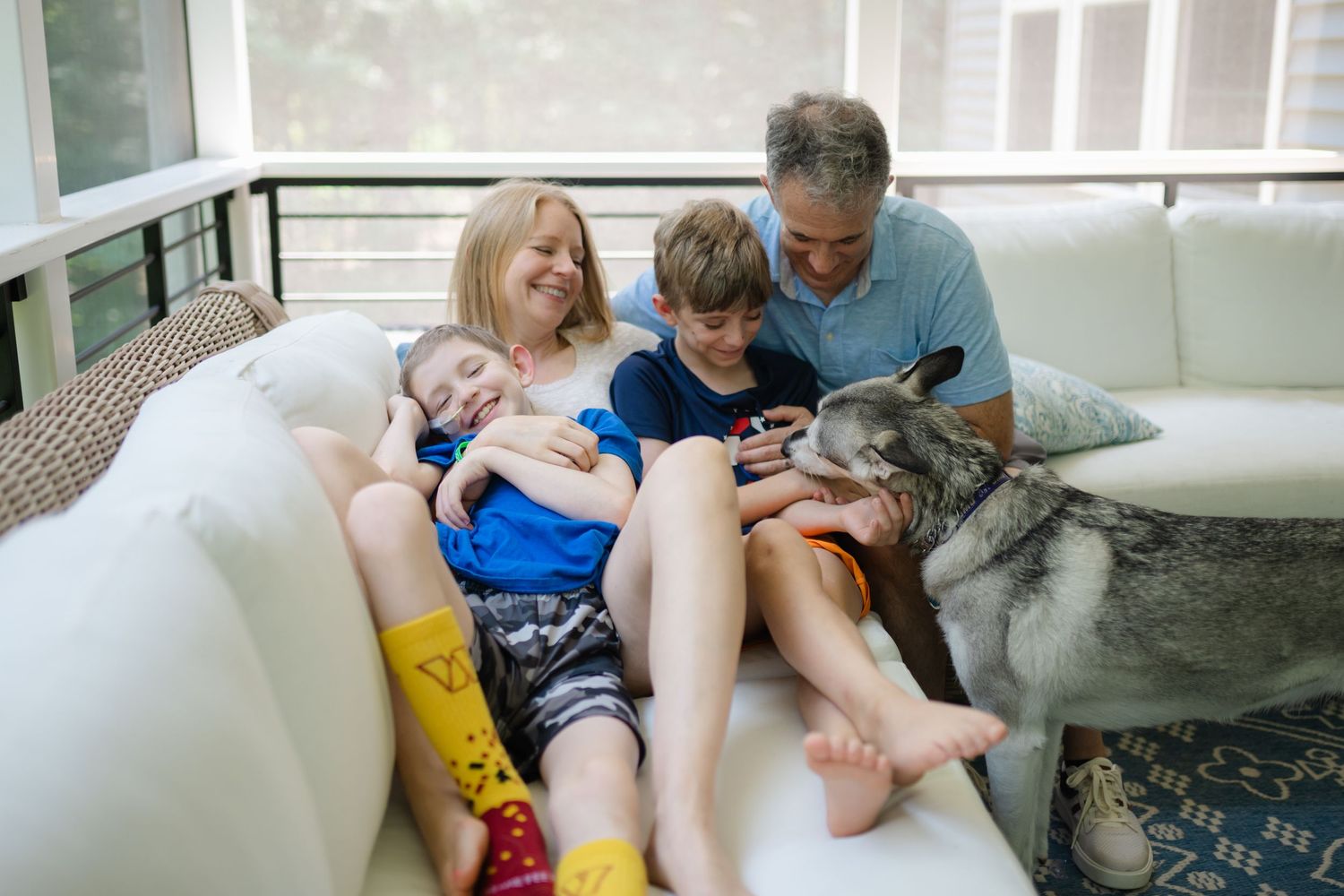 Family of four relaxing on white sofa with husky dog, parents and two children smiling and bonding together indoors.