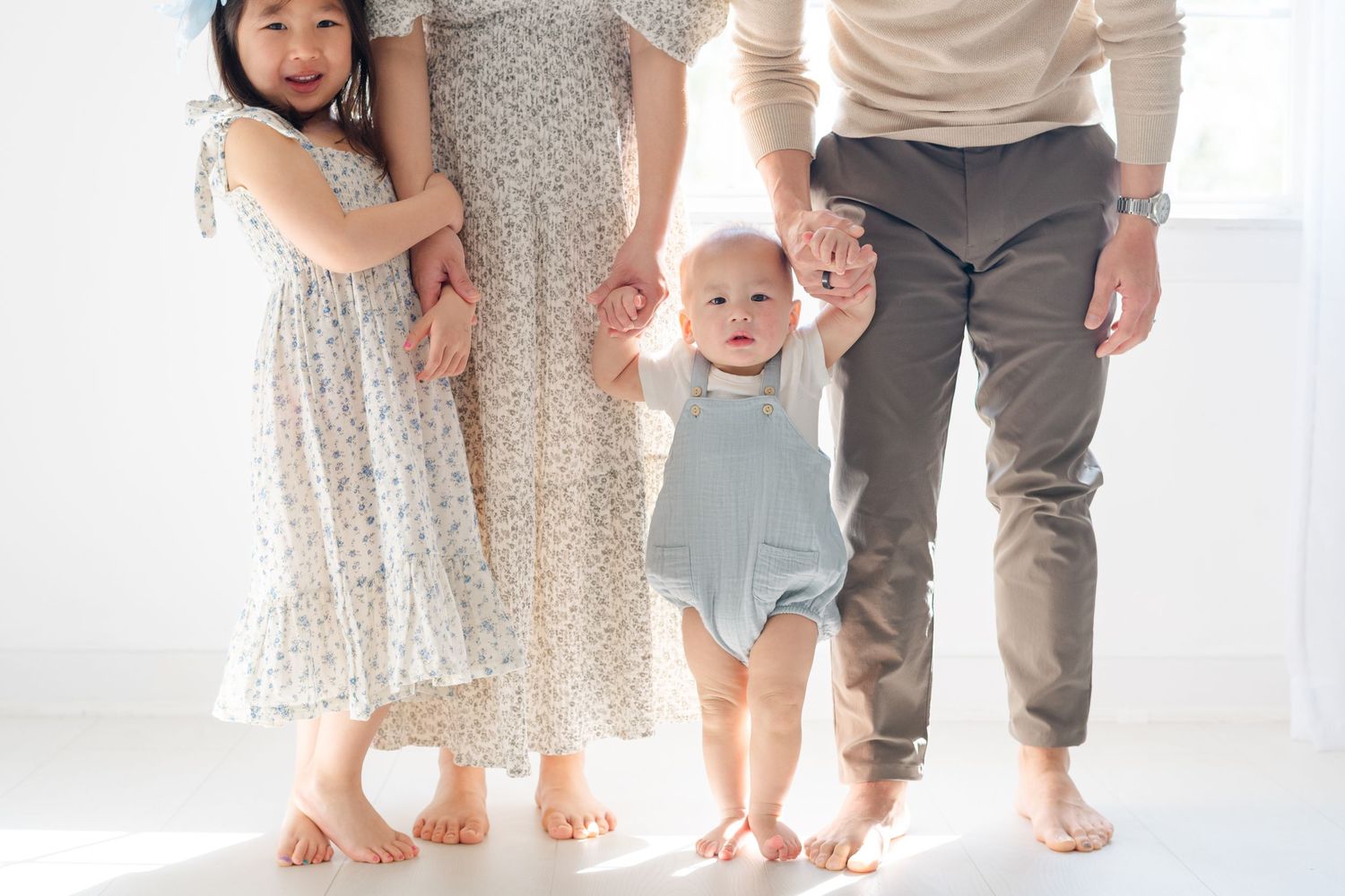 Family of four with toddler taking first steps, parents and young girl holding hands in bright white studio.