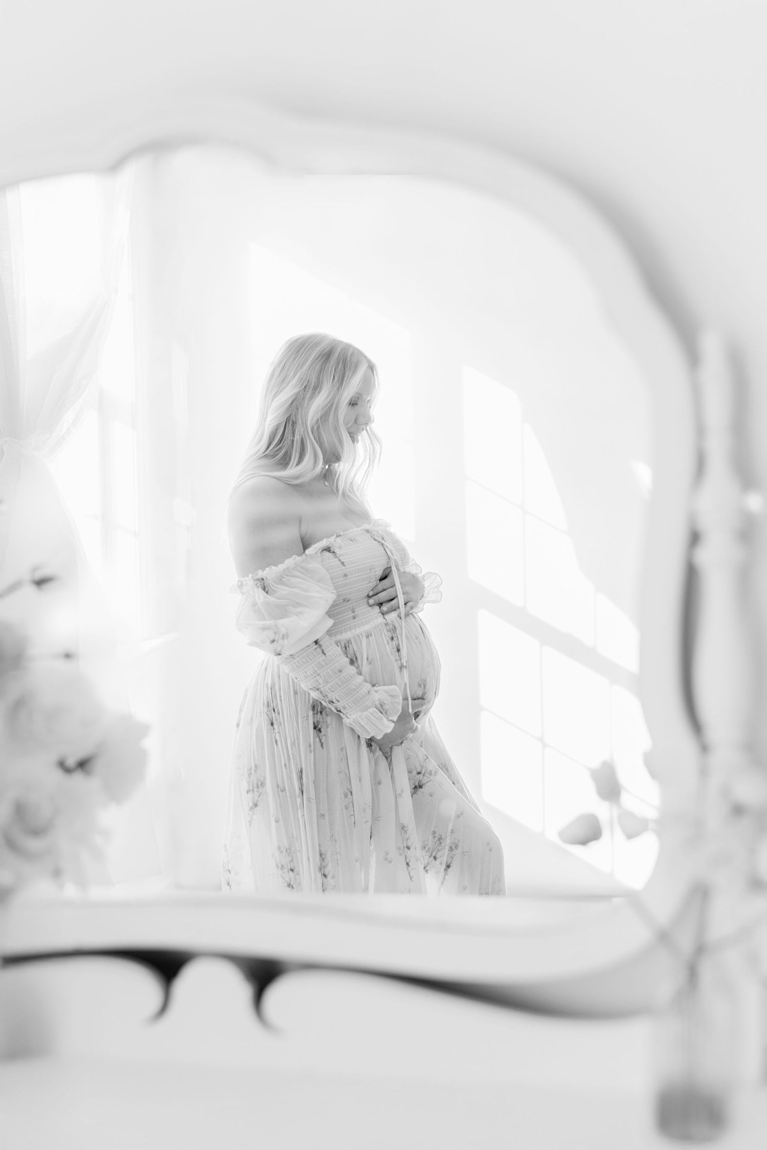 Black and white mirror reflection of a pregnant woman in a floral dress cradling her baby bump for maternity portraits