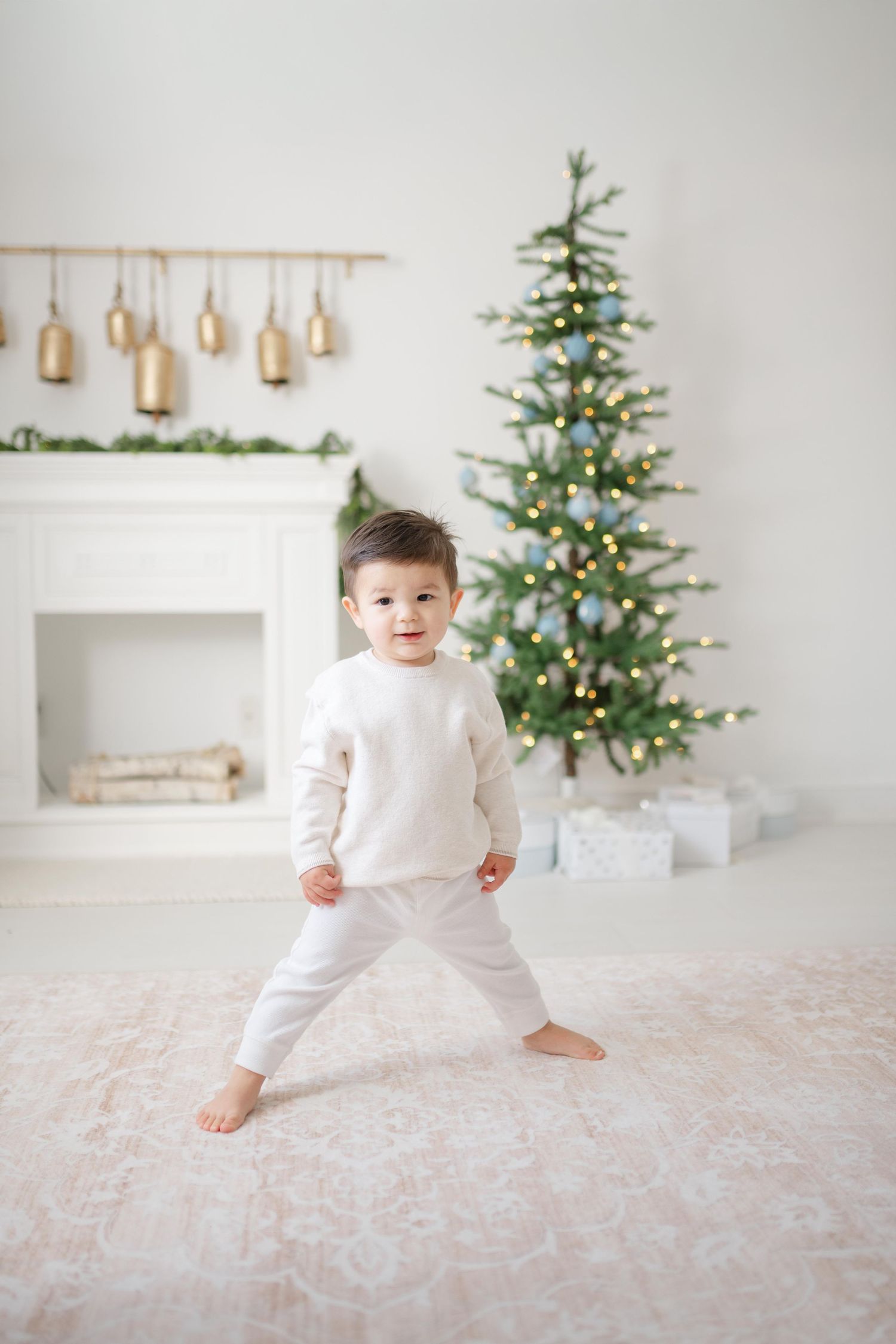 Toddler in white outfit standing barefoot near a decorated Christmas tree with lights and gifts.