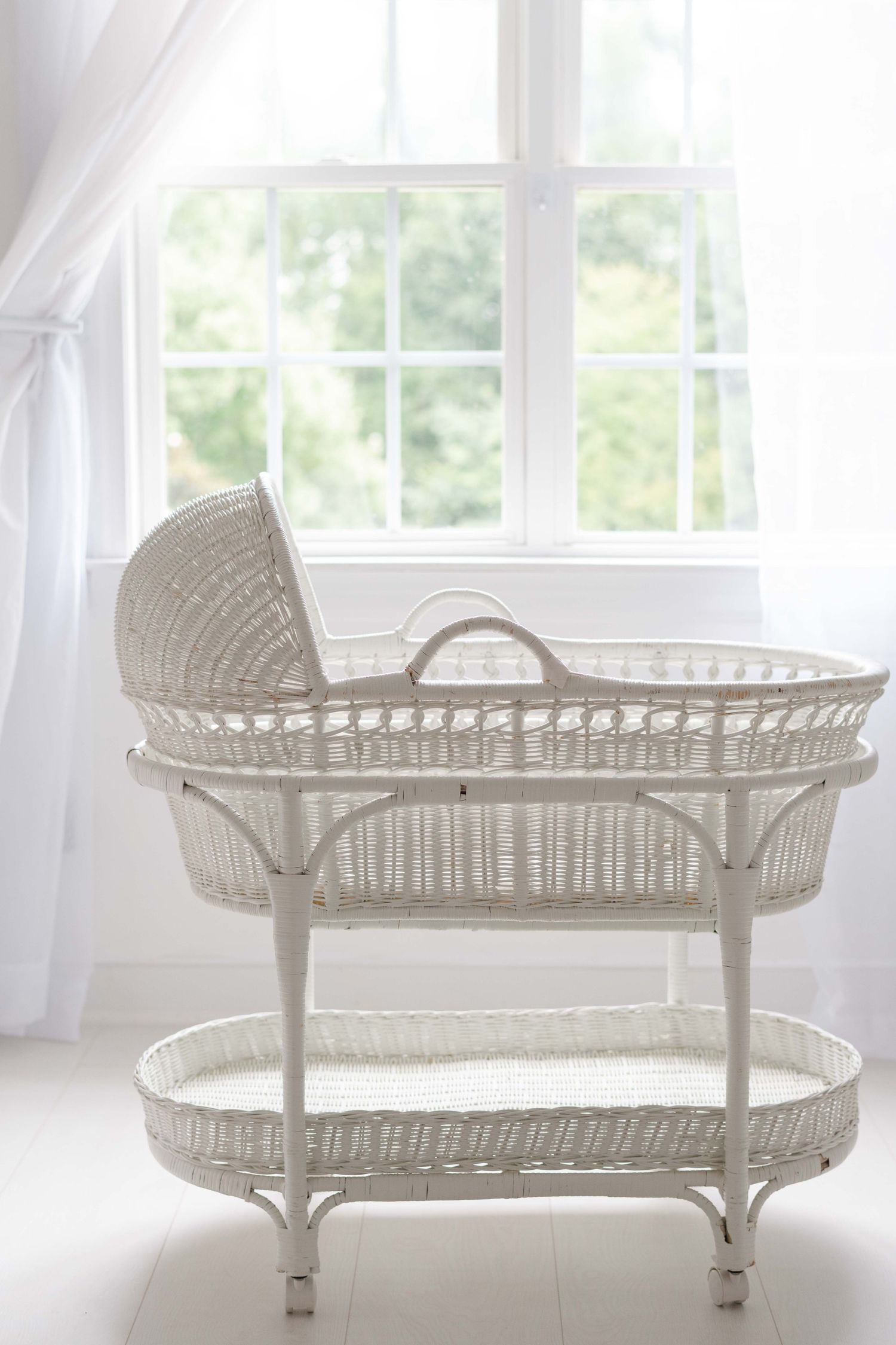 White wicker baby bassinet on stand positioned in front of bright window with sheer curtains in a serene nursery.