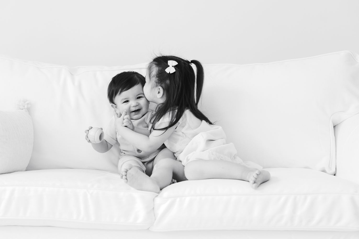 Two children playing together on white couch in black and white photo.