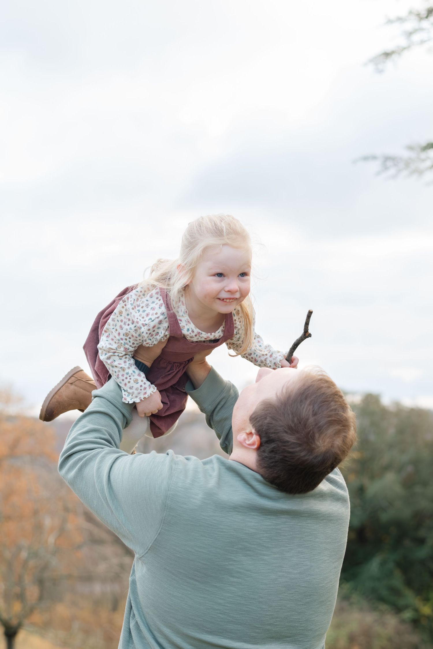 Father lifting smiling blonde toddler girl into the air outdoors during autumn family photo session.