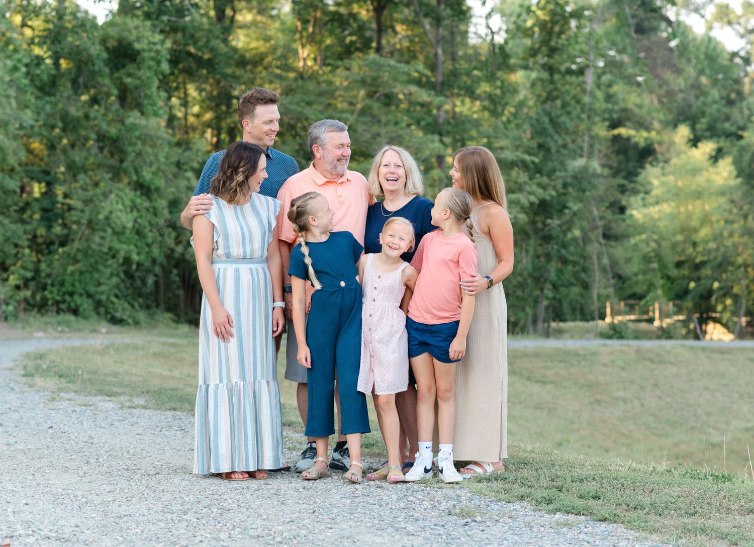 A joyful multigenerational family poses outdoors on a gravel path surrounded by lush green trees in warm summer light.