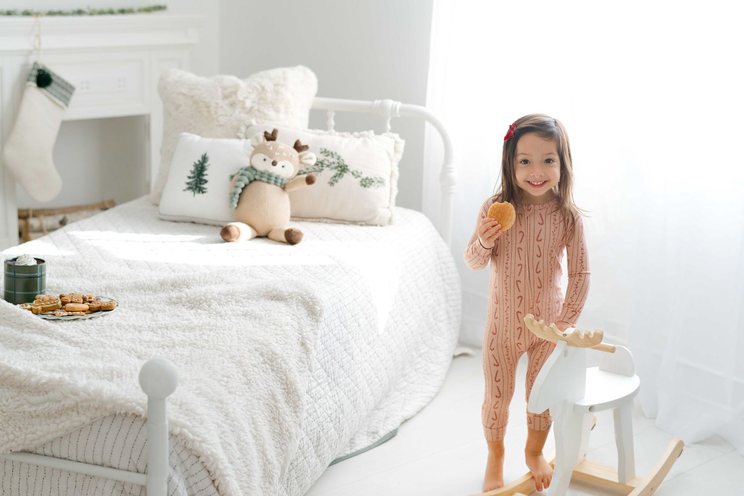 A young girl in a pink knit onesie holds a cookie beside a white bed with holiday pillows and a stuffed reindeer toy.