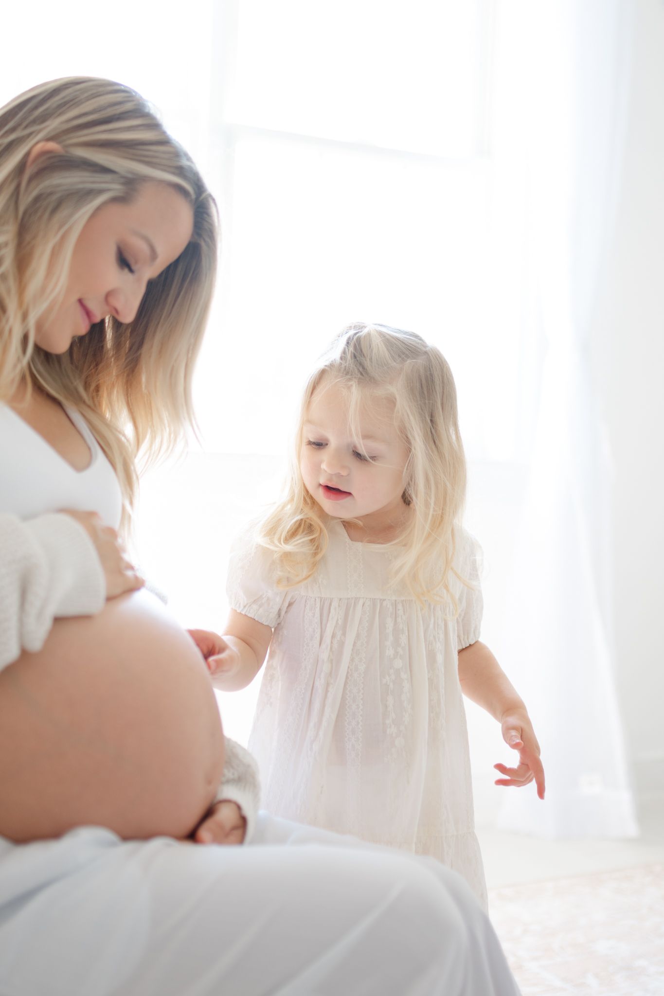 A tender moment between a pregnant woman in white and a small child with blonde hair in a bright sunlit midlothian photography studio
