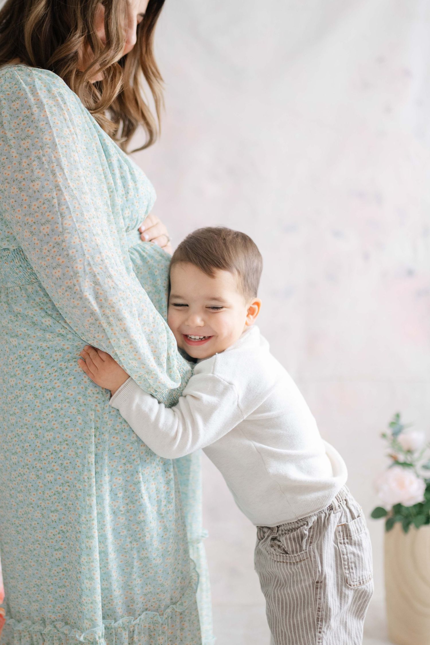 Candid moments between mother in blue dress and toddler in white against light backdrop.