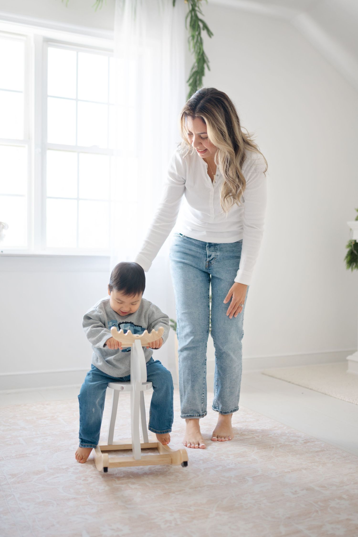 A mother smiles as her toddler plays on a wooden rocking horse toy in a bright, minimalist white room.