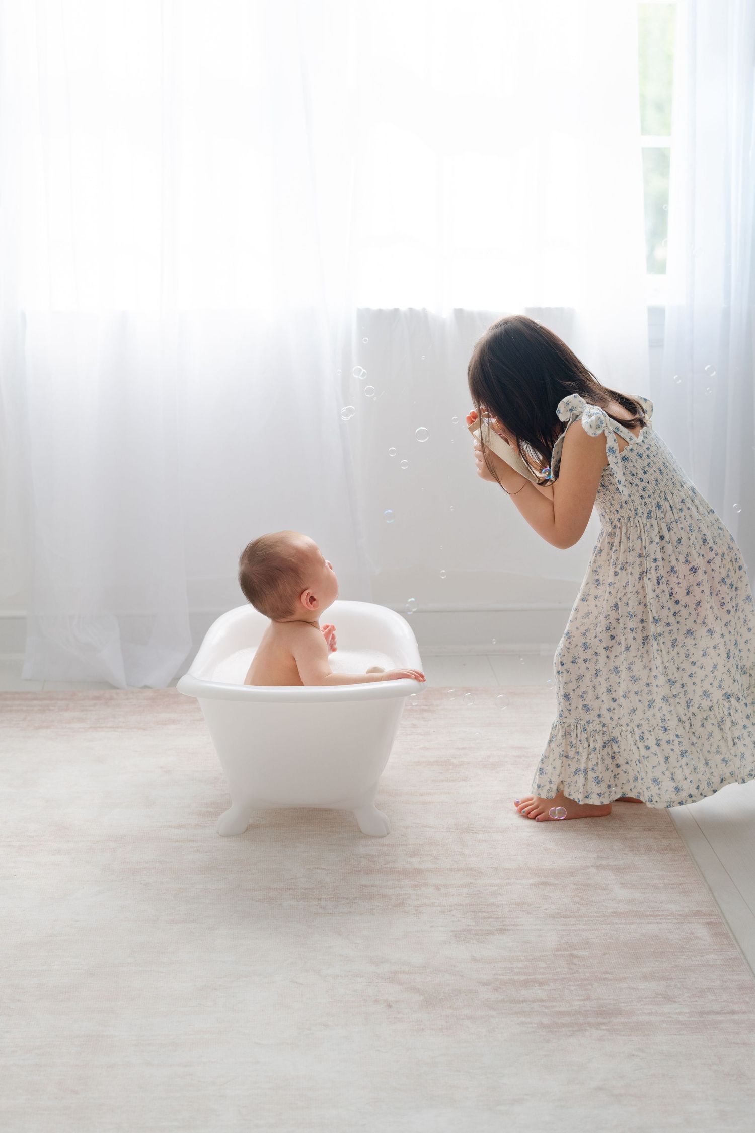 Young girl in floral dress blowing bubbles at smiling baby sitting in small white bathtub indoors.