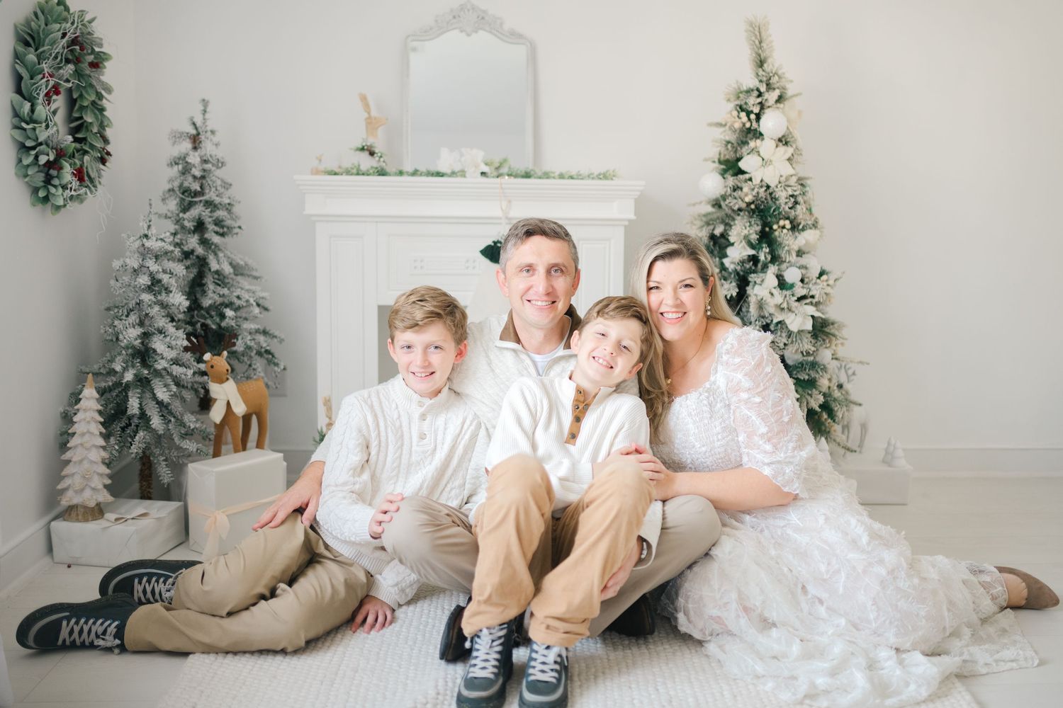 A family in matching white outfits poses together on a couch during a Christmas photoshoot with decorated trees.