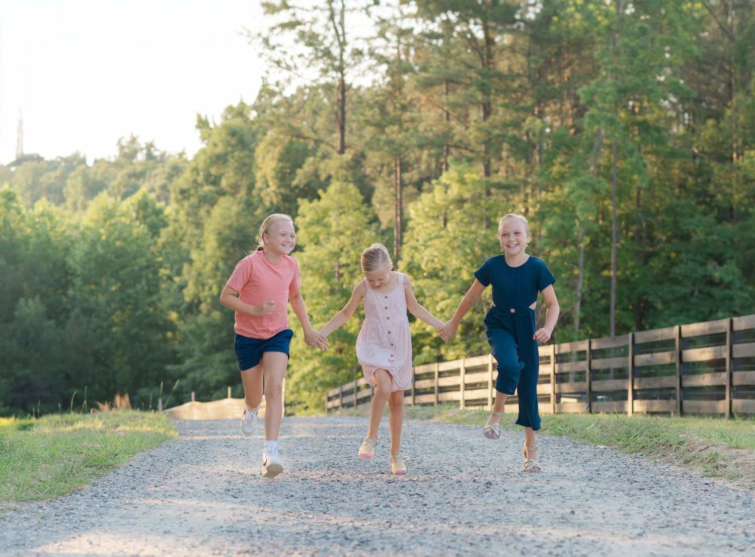 Three children running joyfully on a gravel path surrounded by green trees and a wooden fence.