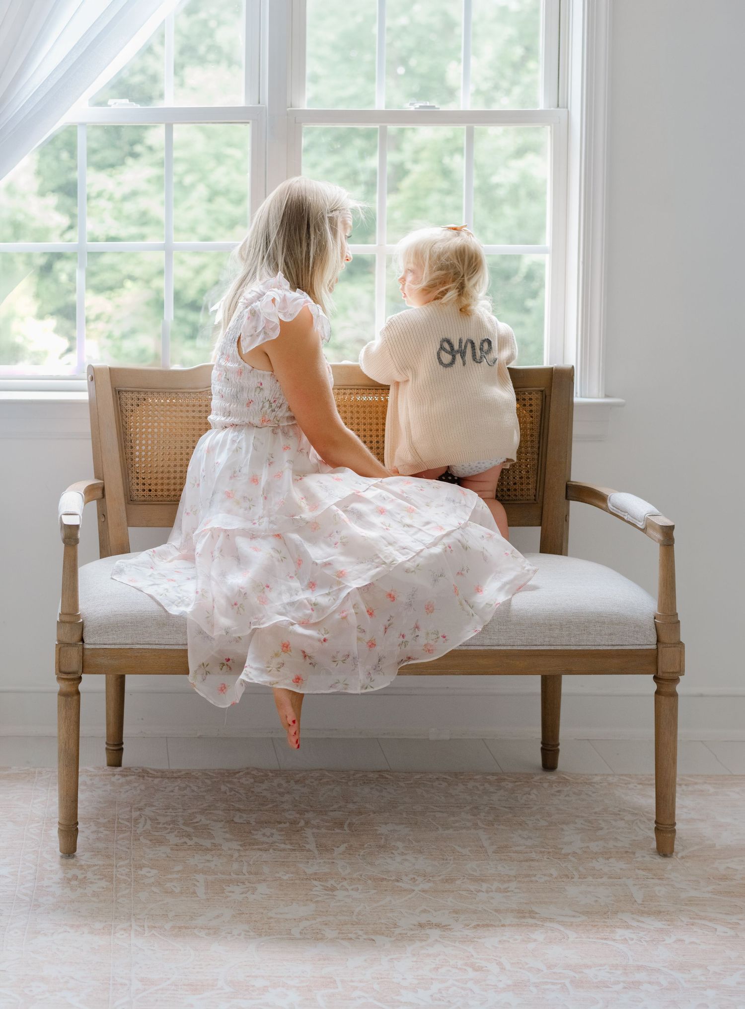 Mother and toddler in floral dress sit on cane bench by window, baby wearing 'one' birthday shirt.