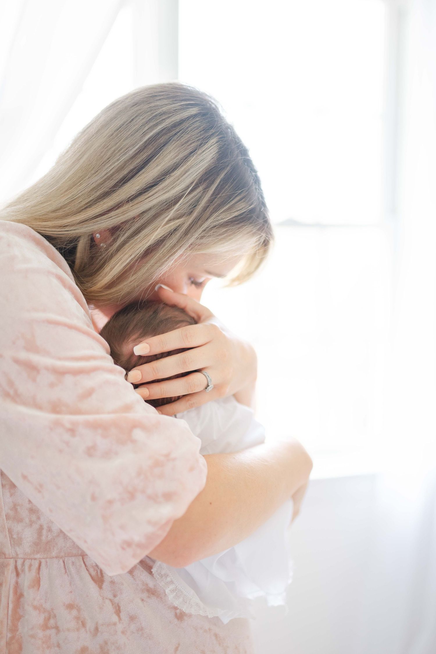 A blonde mother in a pink dress tenderly holds and kisses her newborn baby near a bright window.