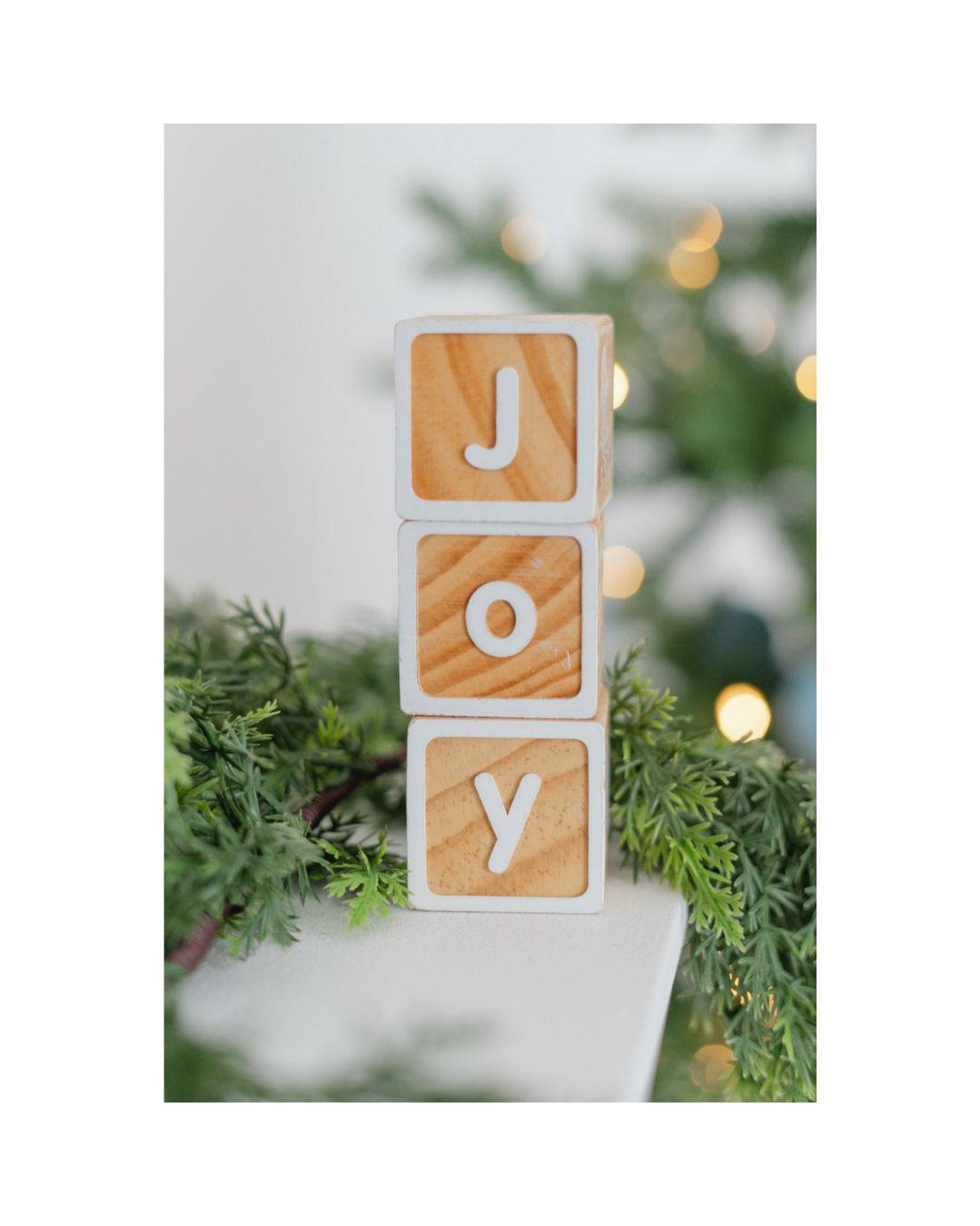 Wooden letter blocks spelling JOY stacked vertically, displayed as a festive Christmas decoration near a green tree.