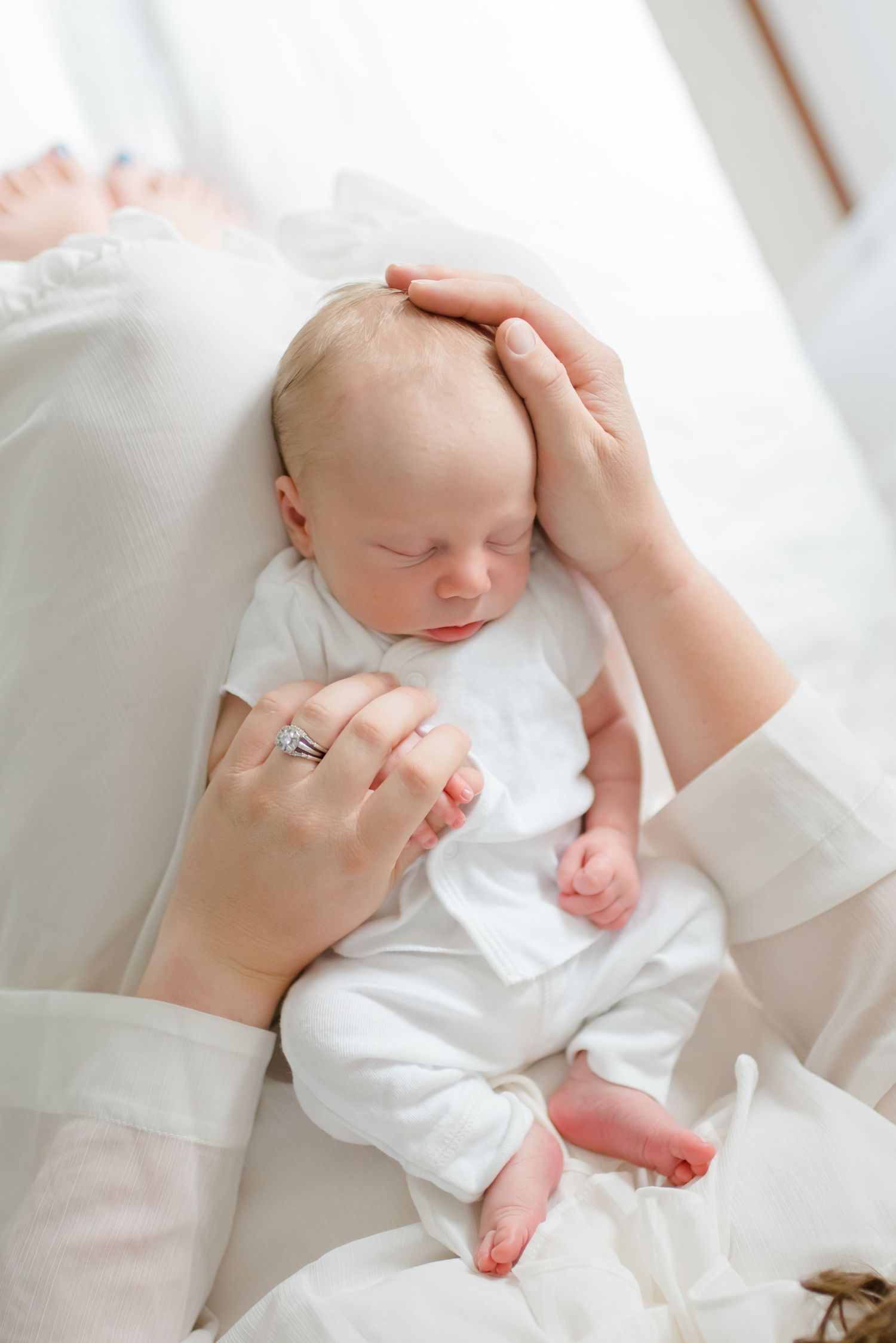 Tender moment of a sleeping newborn in white being gently cradled against soft white bedding.