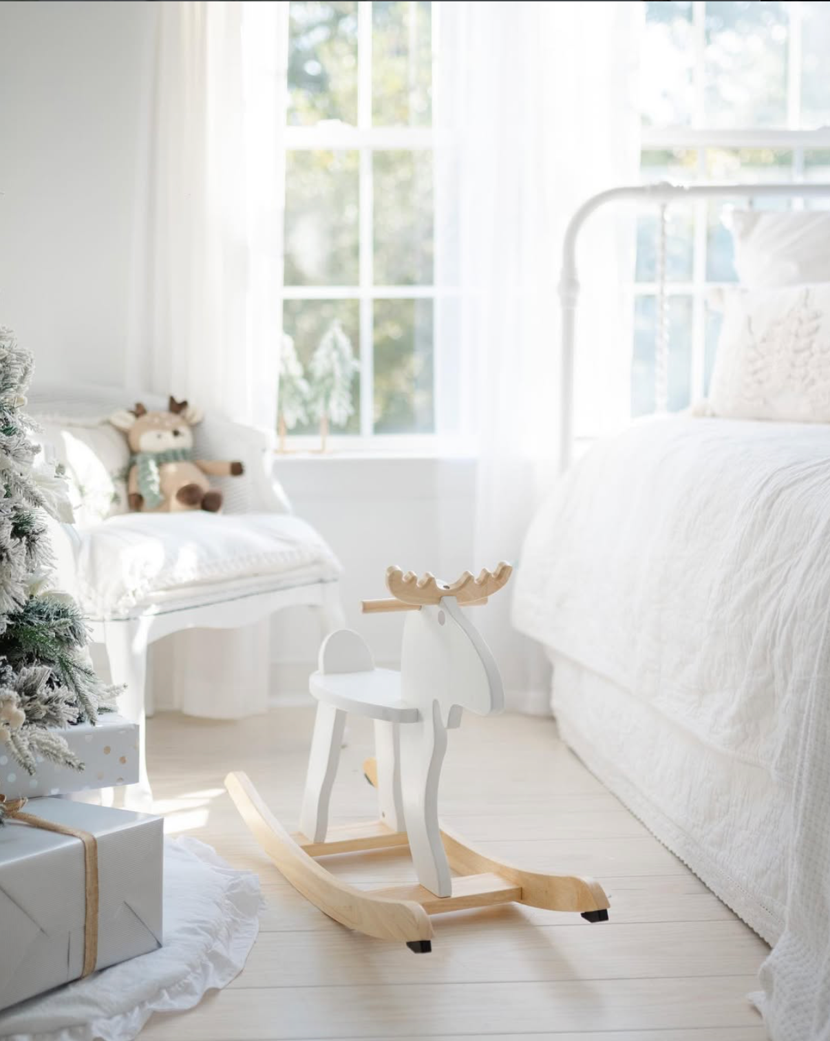 White children's bedroom with wooden rocking moose toy, flocked Christmas tree, wrapped gifts, and bright window.