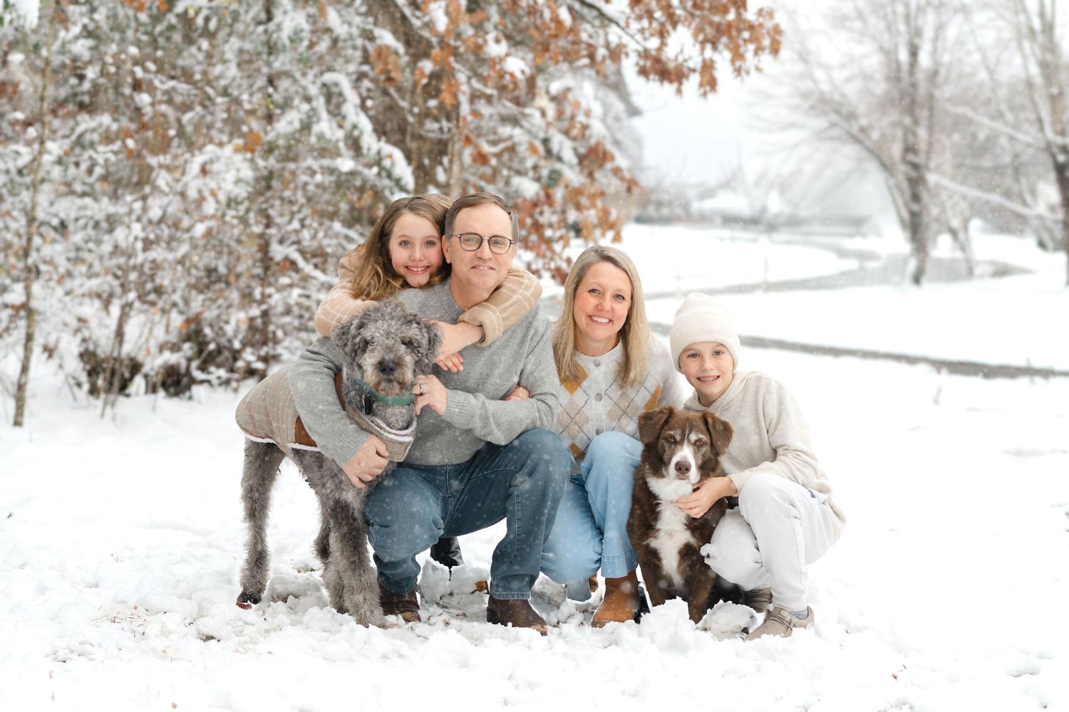 Family of four with two dogs posing happily in a snowy winter landscape surrounded by snow-covered trees.