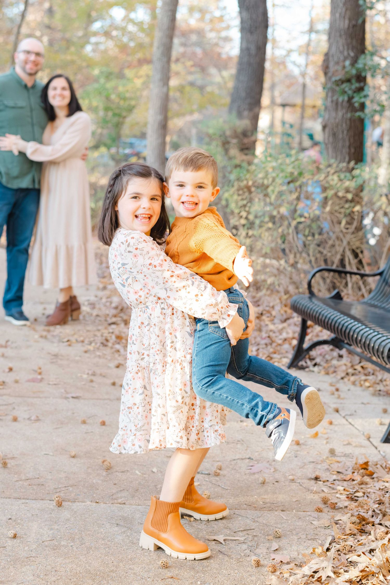 A joyful girl in a floral dress holds her smiling brother outdoors while their parents watch warmly in an autumn during a fall phot session
