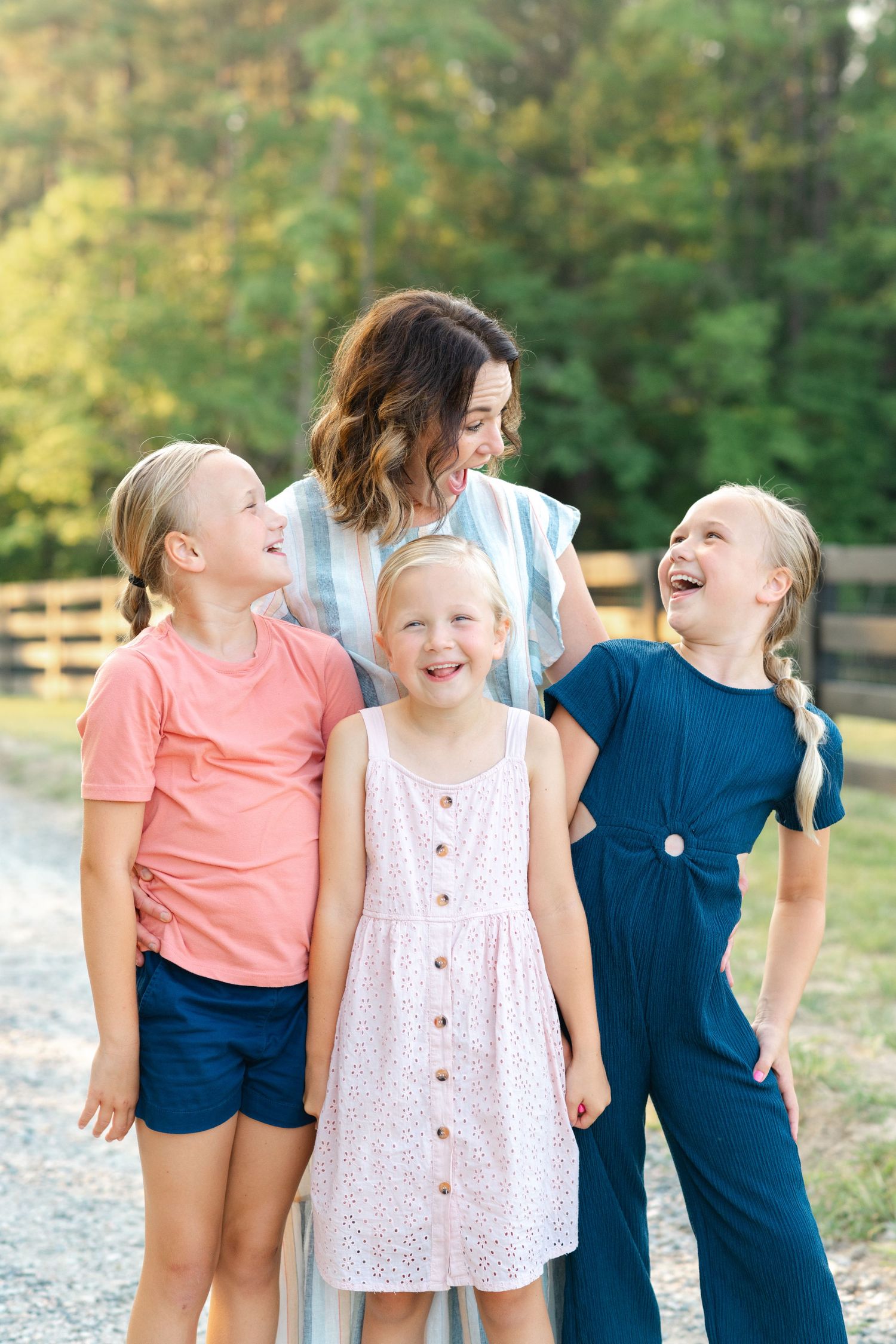 A smiling mom with three laughing blonde girls poses outdoors near a wooden fence surrounded by green trees.