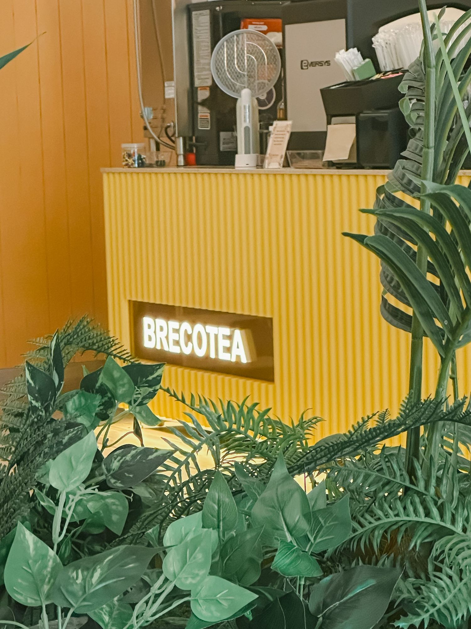 A collection of artificial tropical plants arranged against a yellow wall with a neon sign in the background.