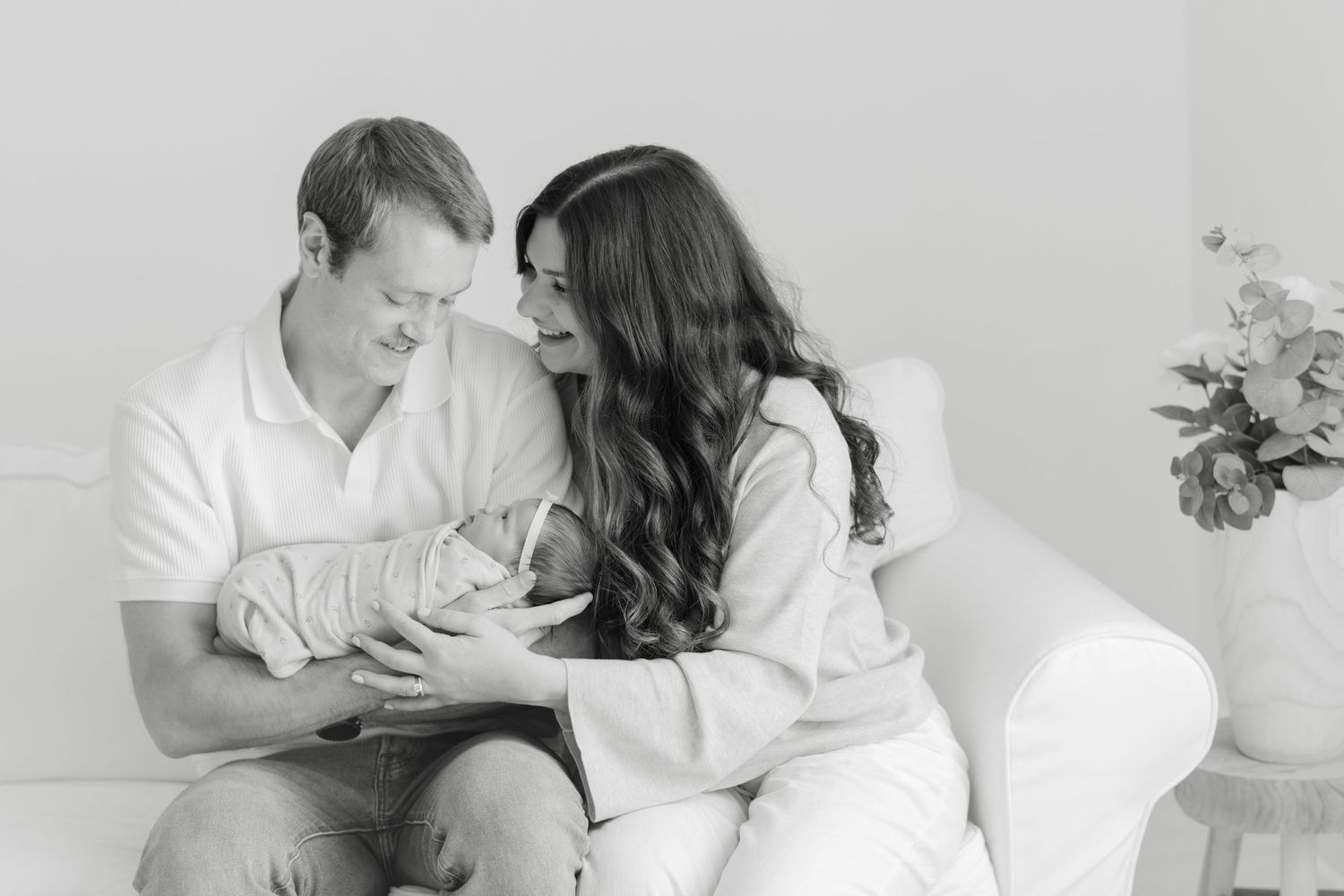 Black and white photo of a couple sitting on a couch, lovingly gazing at their newborn baby.