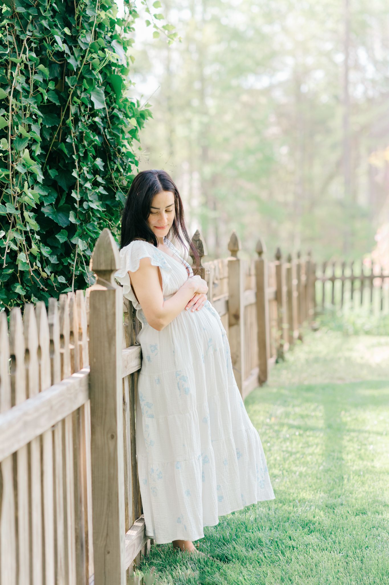 Woman in white dress standing by wooden fence in garden setting.