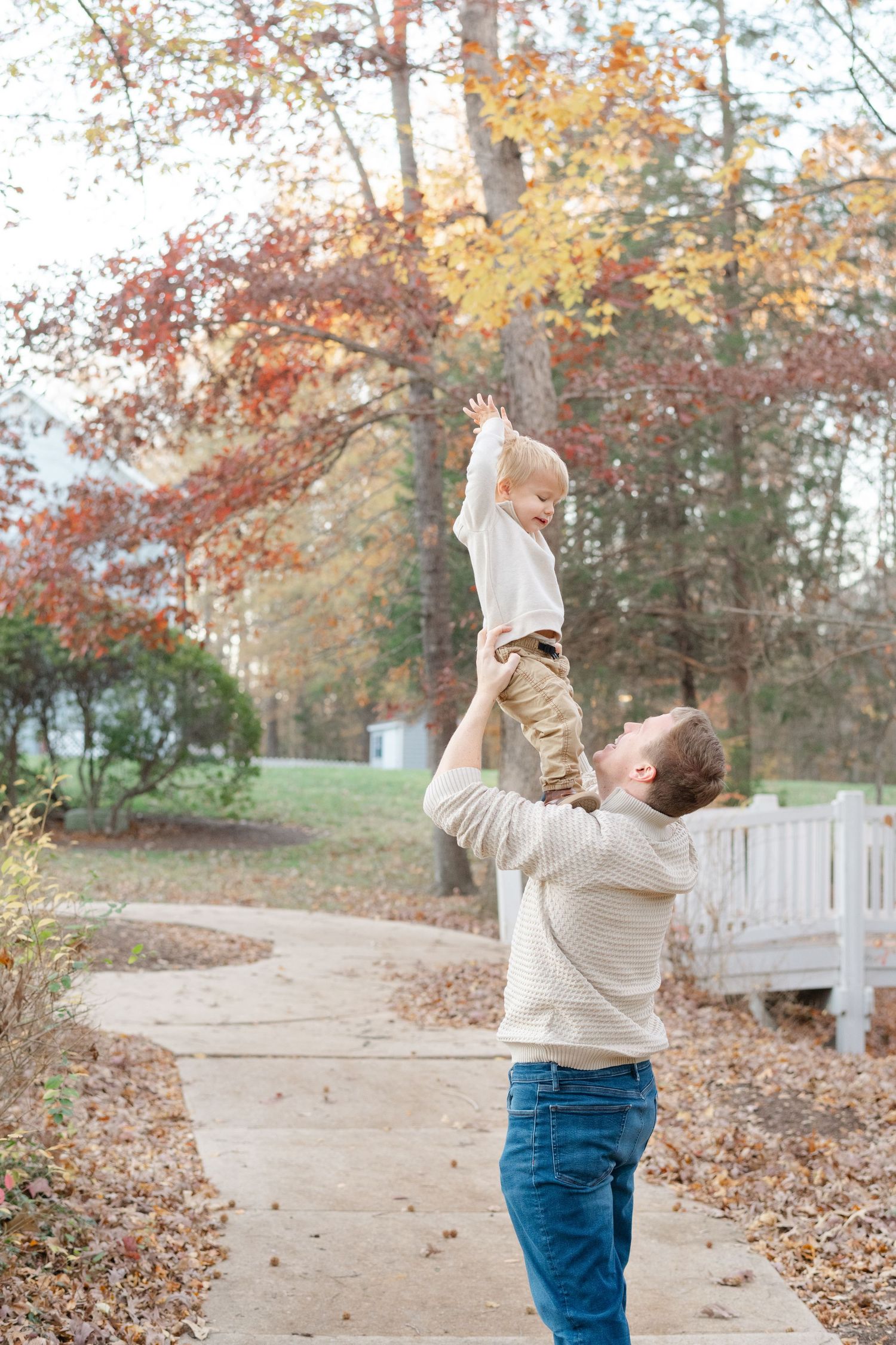 Father joyfully tosses toddler in the air on a tree-lined autumn path, surrounded by colorful fall foliage.