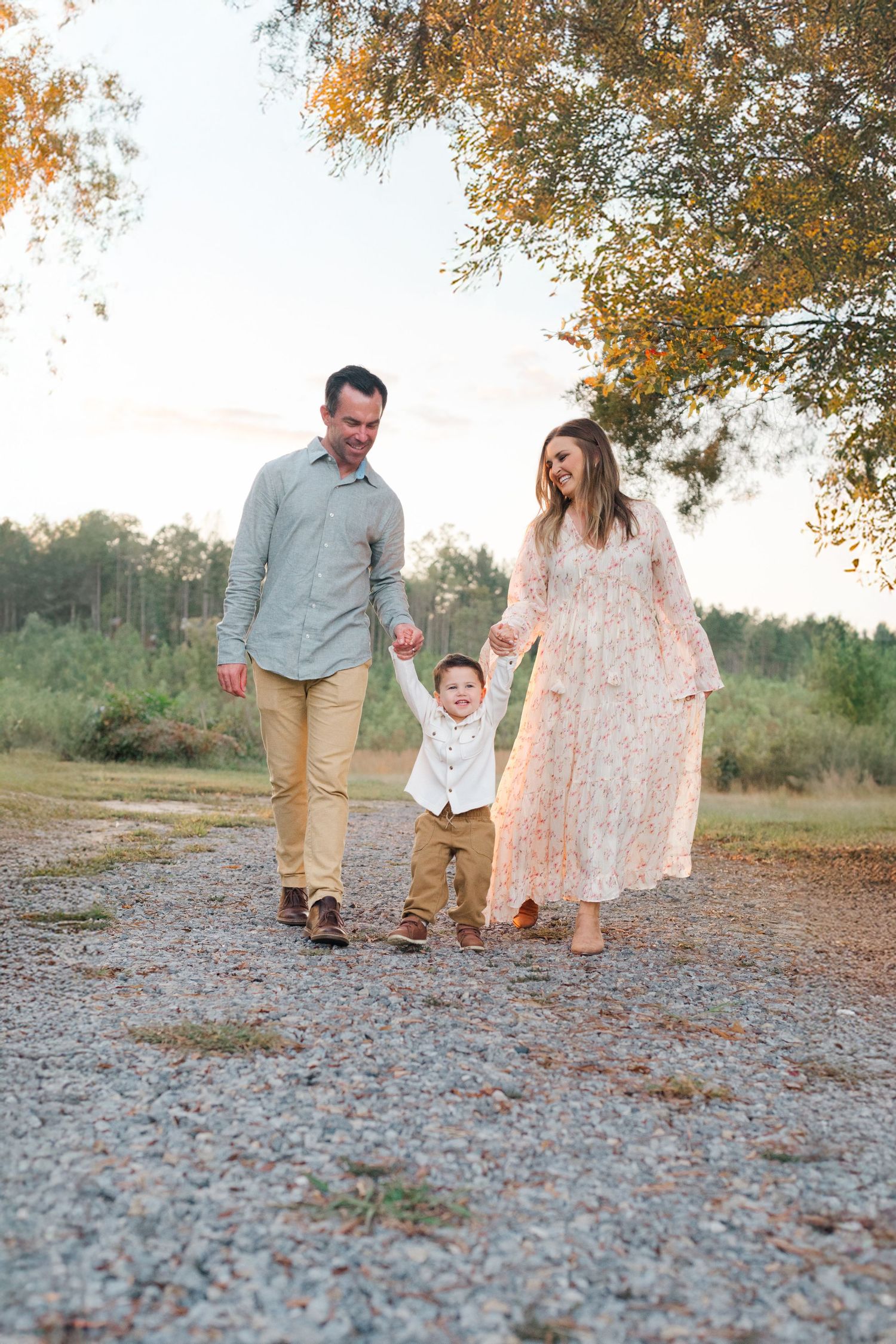 A happy family of three walks hand-in-hand on a gravel path surrounded by autumn trees at golden hour.