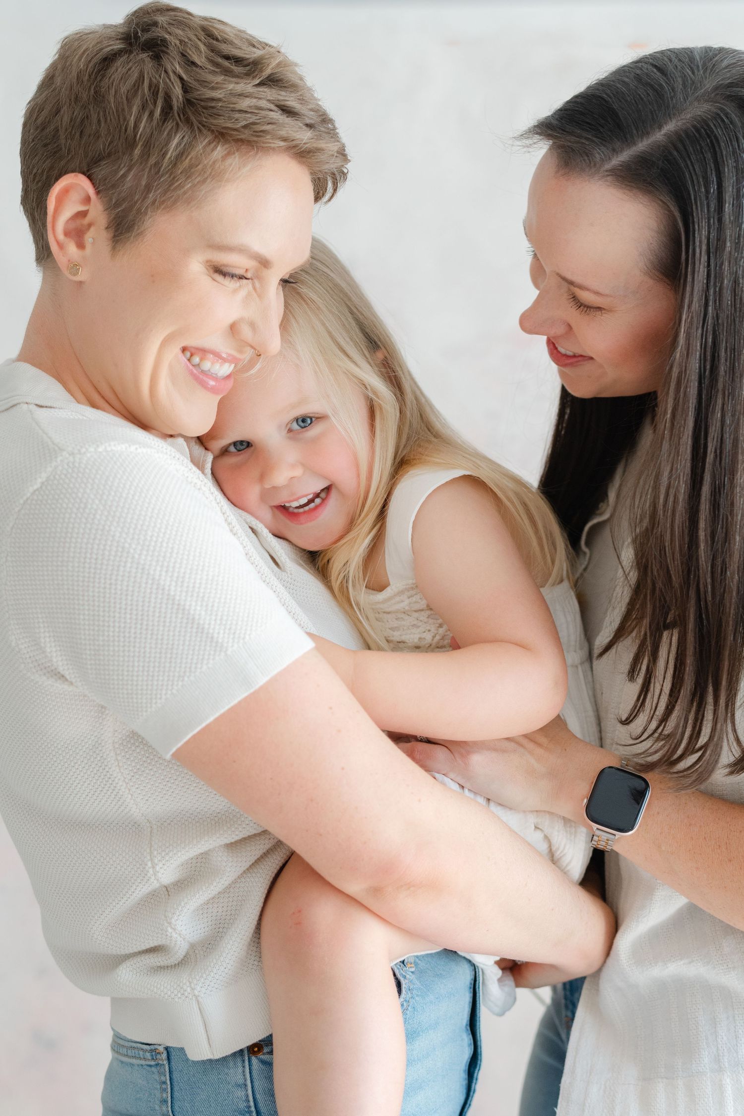 Two women smiling and laughing with a young blonde girl in a bright, cheerful family portrait.