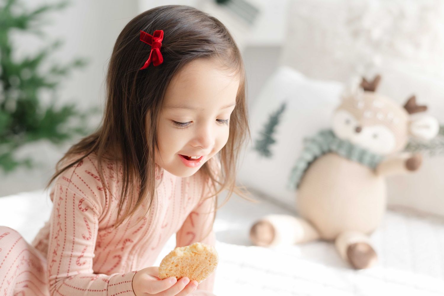 A happy little girl in pink pajamas holds a cookie near a Christmas tree and festive reindeer decor.