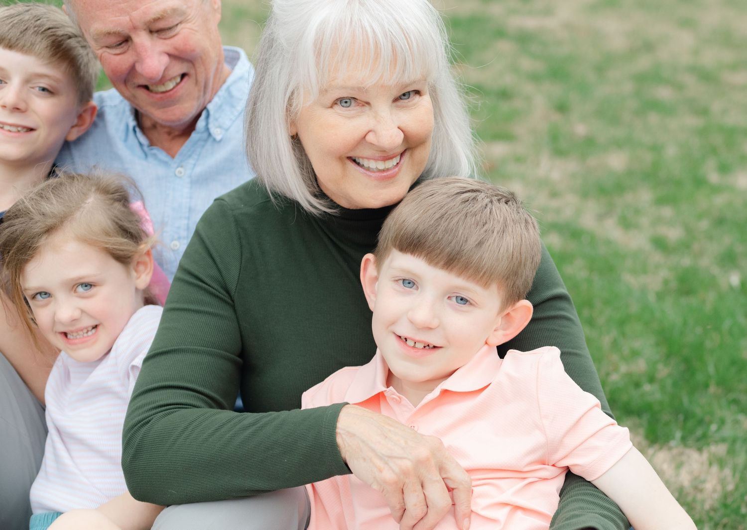 Grandparents smiling outdoors with three grandchildren, enjoying a warm family moment together on green grass.