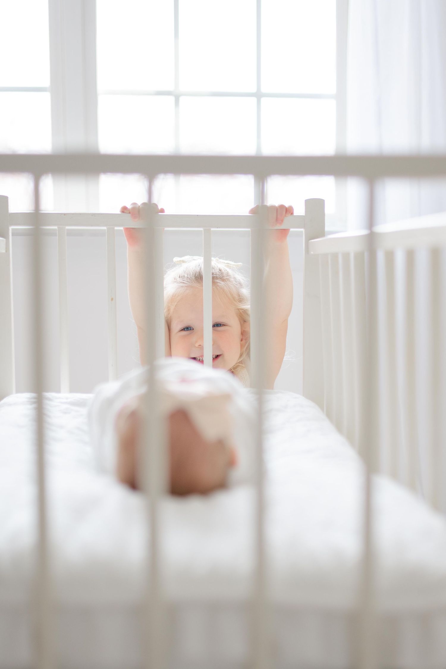 A smiling baby in white clothing lies in a white crib, reaching up to hold the crib rails in a bright, airy room.