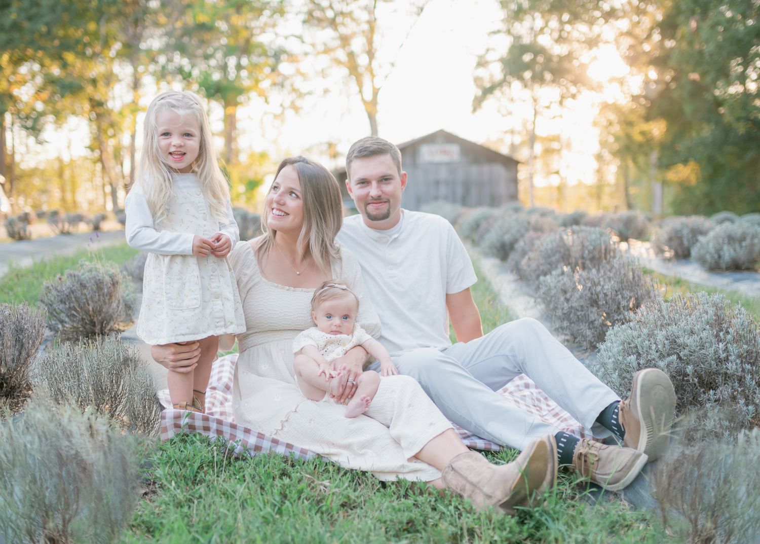 Family poses together on grassy hill during sunset with rustic barn visible in background.