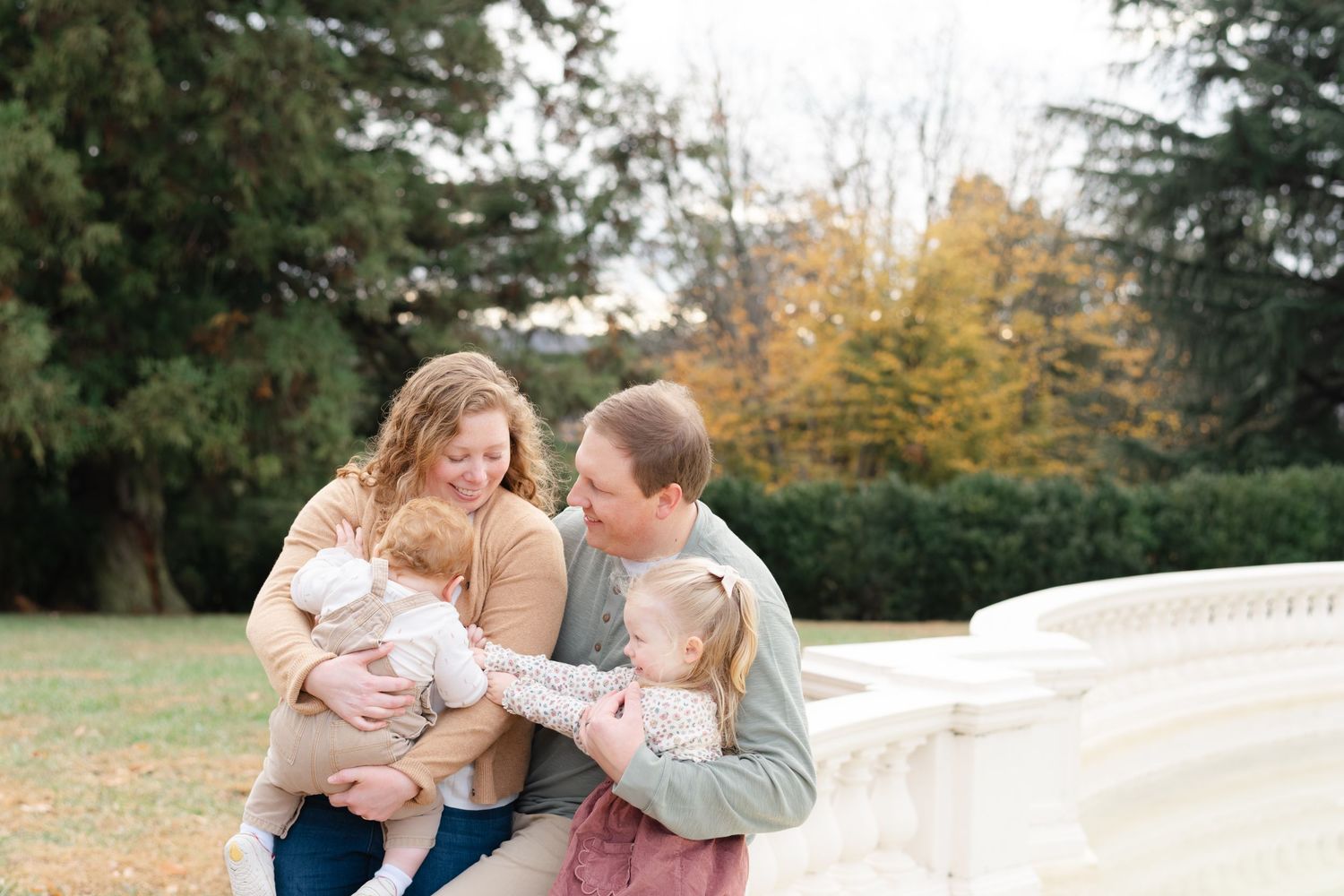 Family of four sharing a joyful moment outdoors in autumn, parents holding two young children near a stone balustrade.