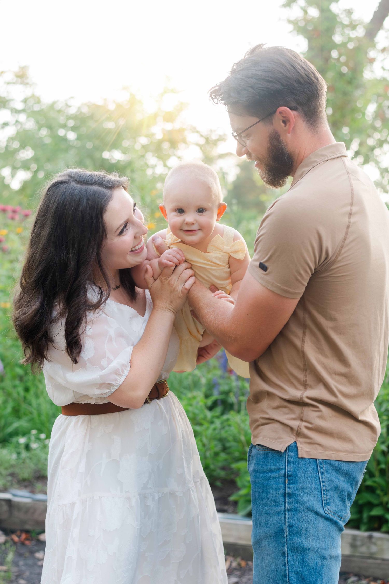 A young family with a bearded father, dark-haired mother in white dress, and smiling baby in yellow outfit pose outdoors.