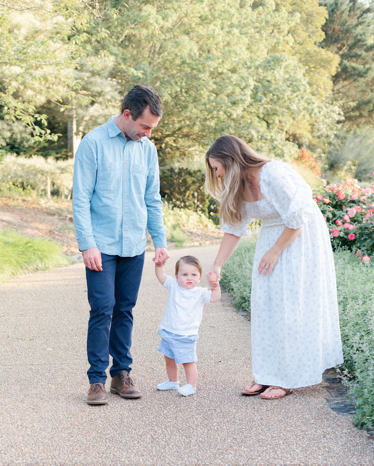 Family helping toddler take first steps on garden path.