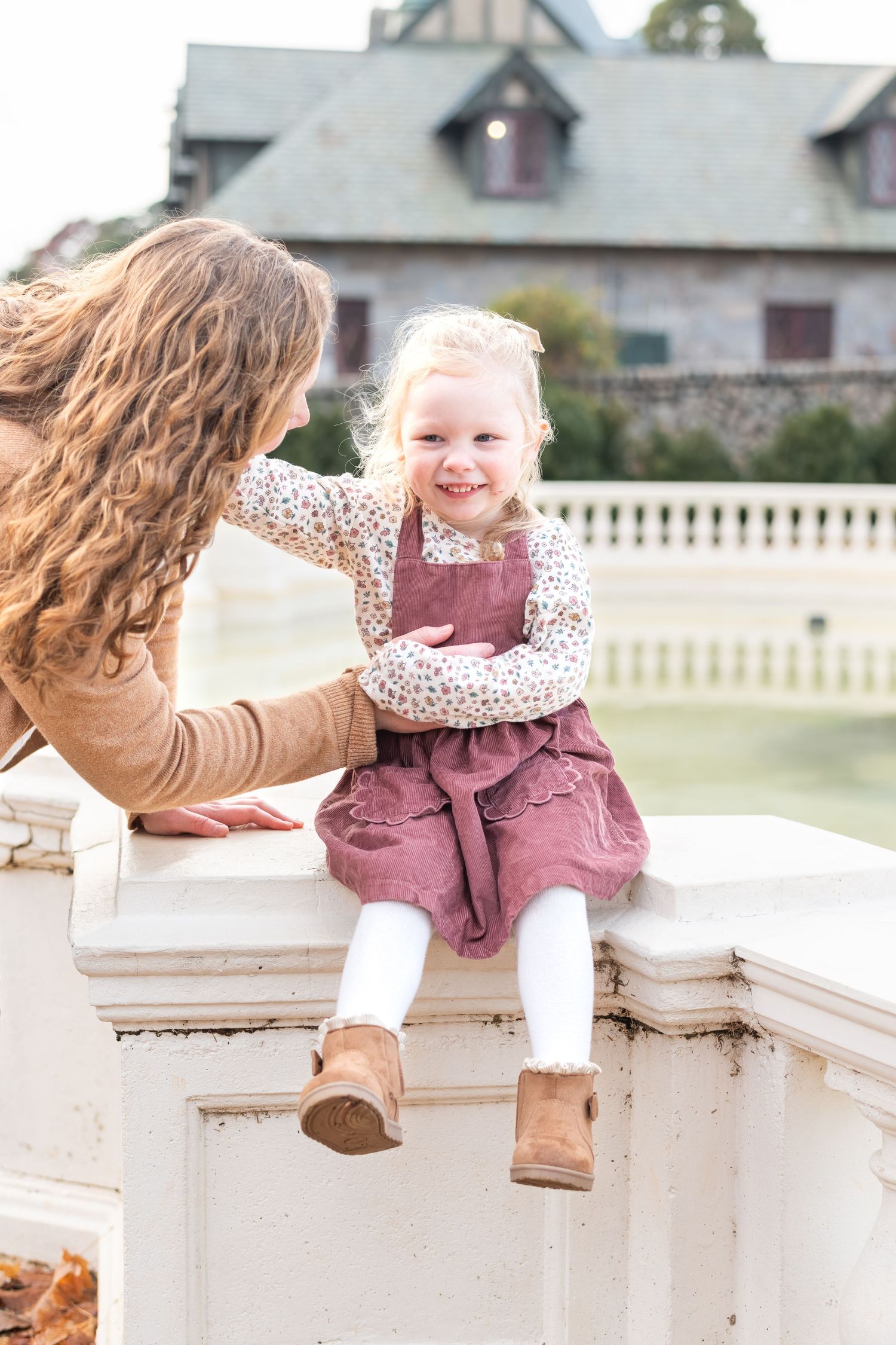 A mother interacts with her smiling toddler girl in a pink dress seated on a white stone wall outdoors.