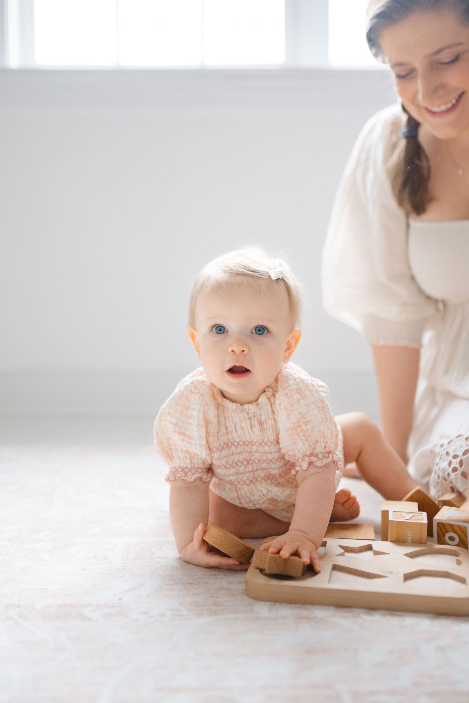 Blue-eyed baby girl in pink smocked romper plays with wooden toys on floor while smiling mother watches nearby.