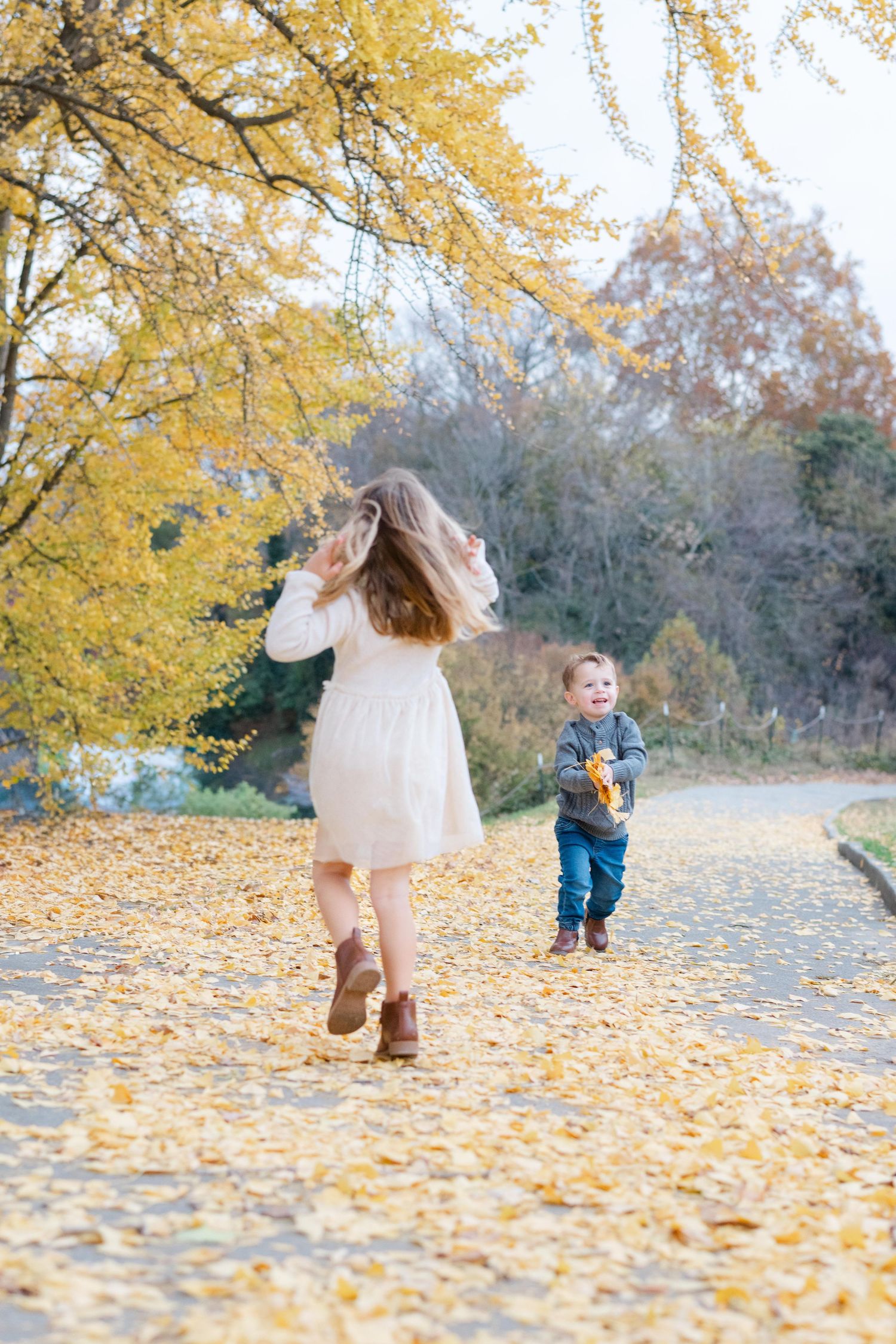 Two children playing on a leaf-covered path surrounded by golden autumn foliage in a park.