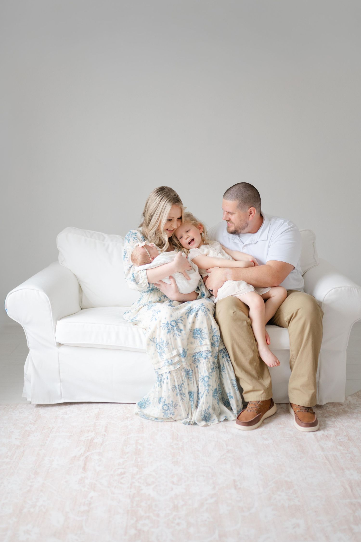 A family in white and beige clothing sits together on a white couch in a minimalist Midlothian studio setting.