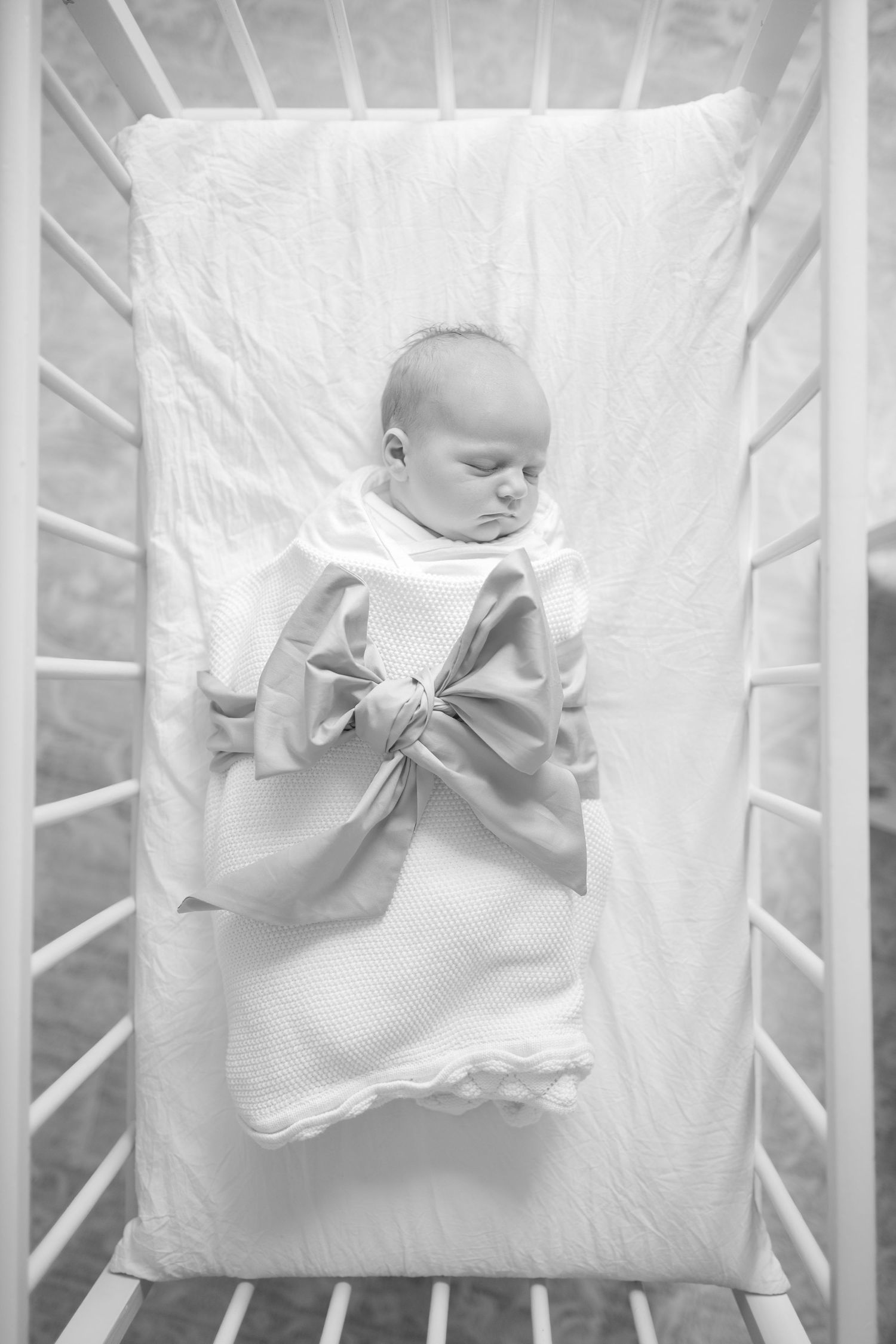 Black and white photo of a sleeping newborn baby wrapped in a blanket with a bow, lying in a white crib.
